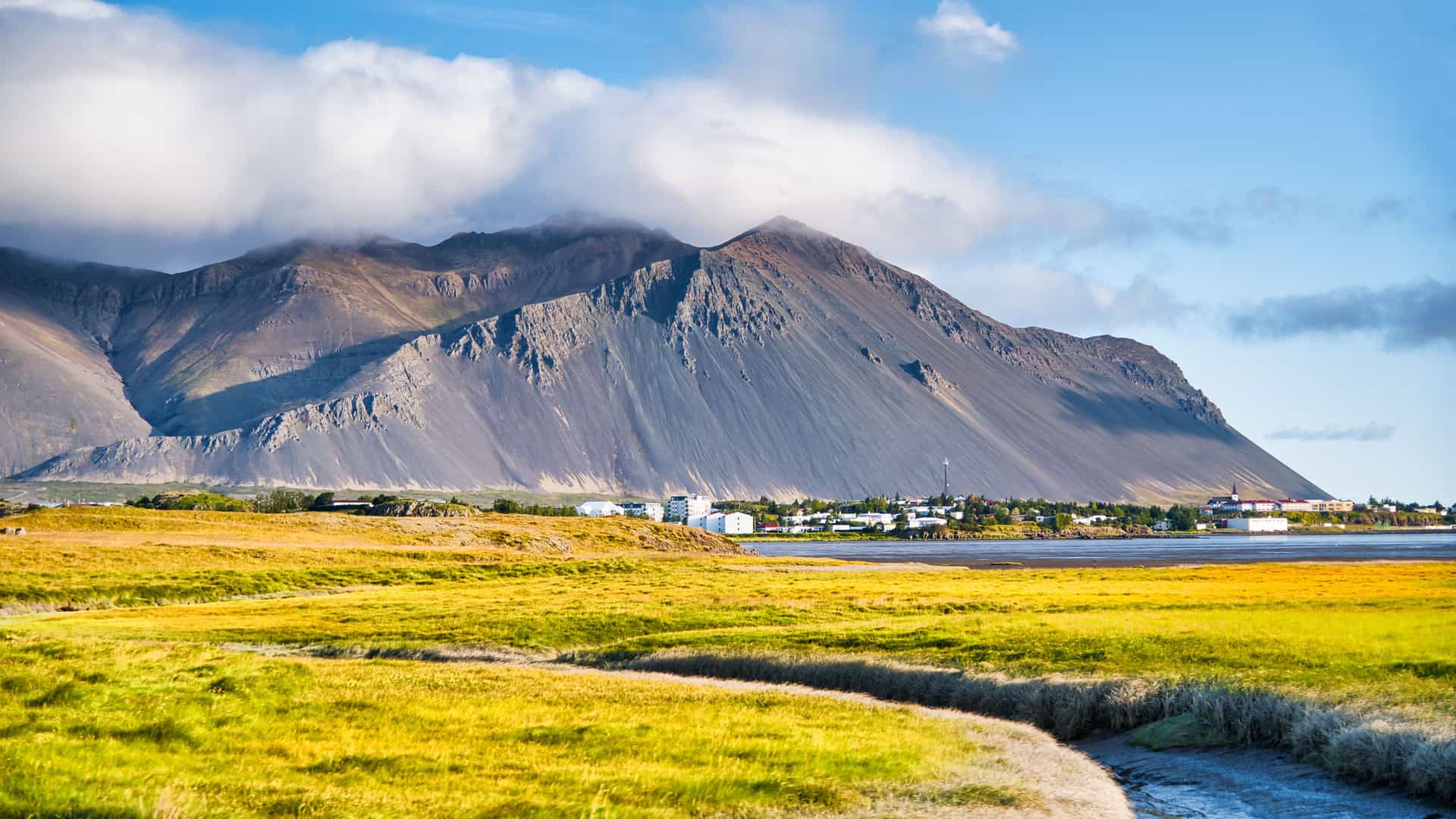 A scenic view of the town of Borgarnes, Iceland, nestled on a flat shoreline. The town's small white and red-roofed buildings are dwarfed by a massive, steep, and rugged mountain range looming behind it. In the foreground, a vibrant field of golden-yellow grass and a small stream lead toward the town, with a mix of blue sky and white clouds overhead.
