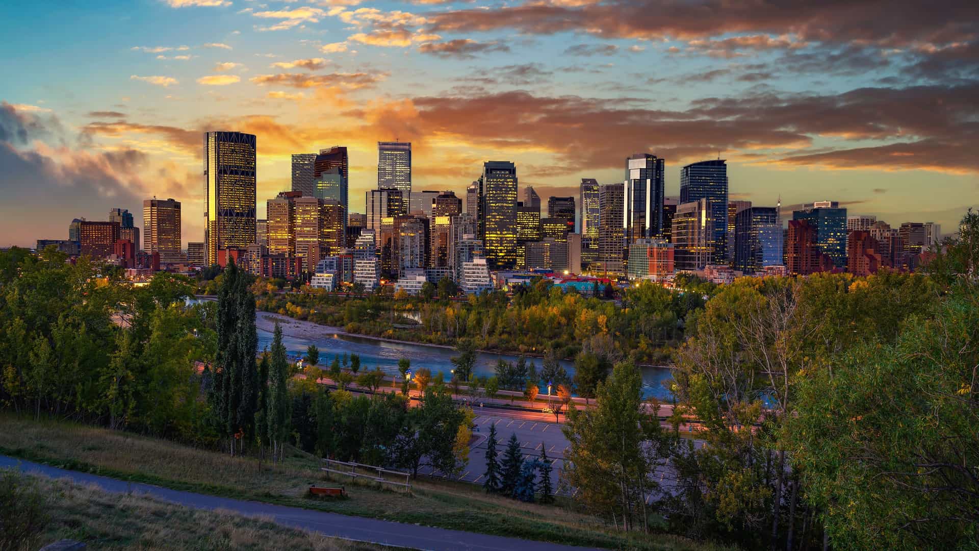 A panoramic, elevated view of the Calgary, Alberta skyline at sunset, with a collection of modern, illuminated skyscrapers rising behind the Bow River. The foreground is a lush, green park with trees and a walking path, all set under a dramatic sky with vibrant orange, yellow, and blue clouds.