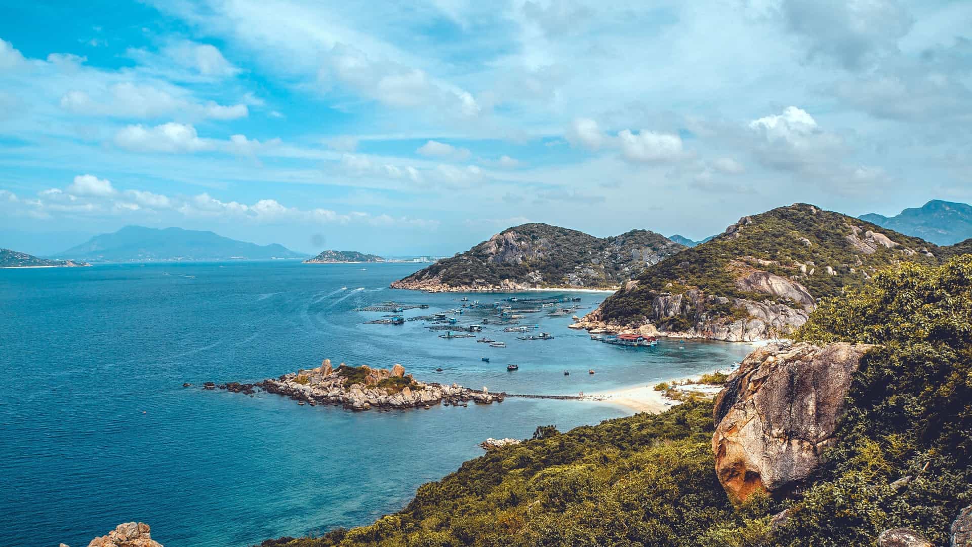 A breathtaking panoramic view of Cam Ranh Bay, with its turquoise waters, lush green mountains, and traditional fishing boats in a serene cove under a beautiful blue sky.