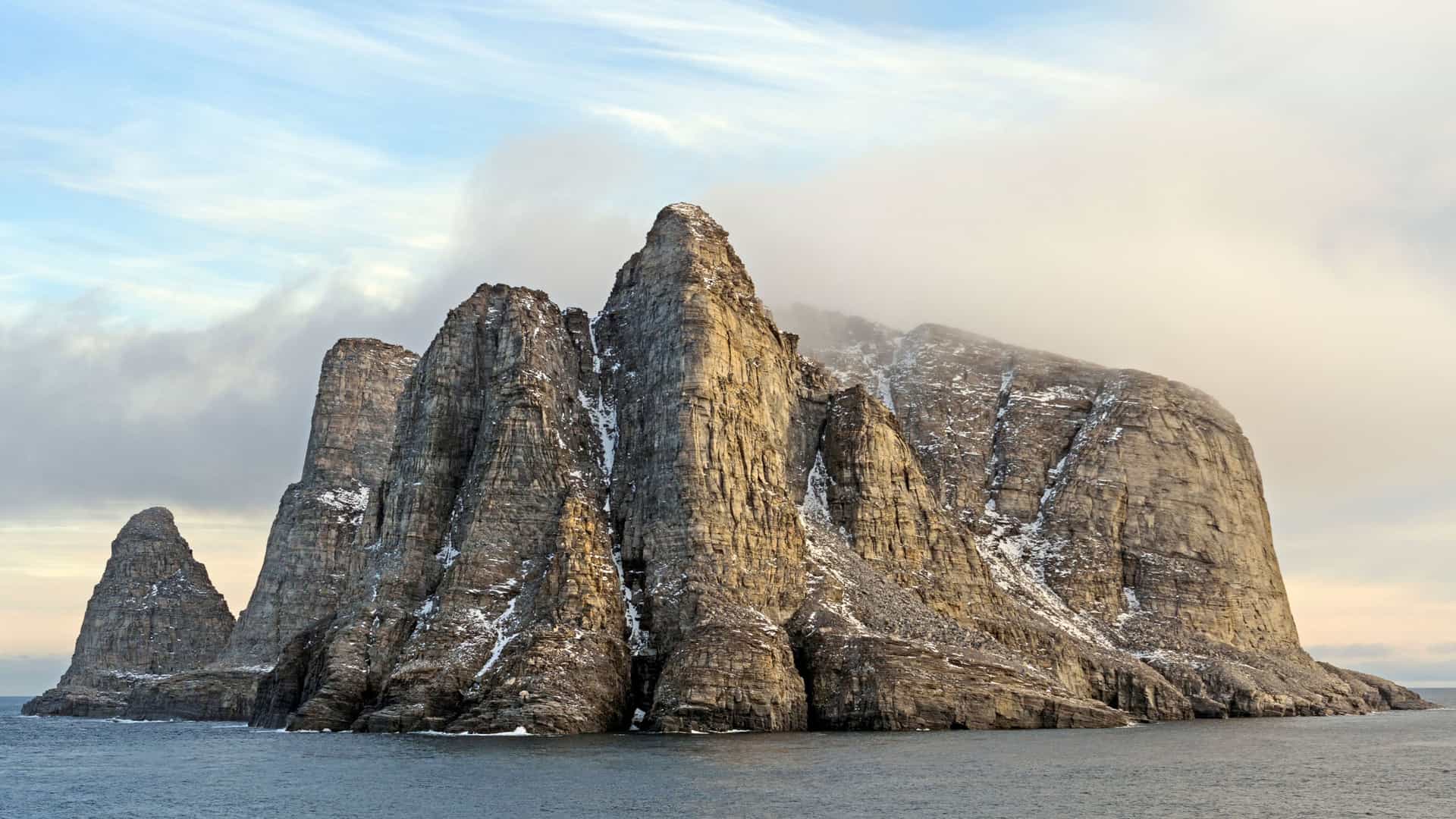 A stunning view of the dramatic, rocky cliffs and sea stacks of Cape Dyer in Nunavut, Canada, a remote Arctic landscape rising from the water with a cloudy sky.