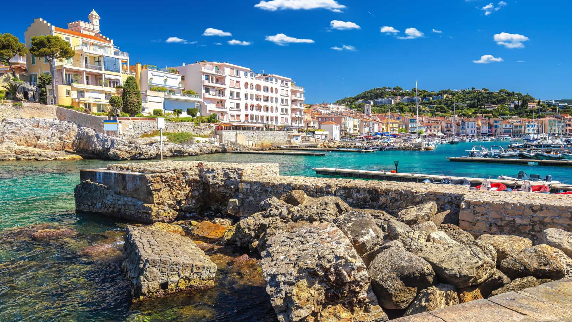 A picturesque view of the charming fishing port of Cassis, France, with colorful buildings and boats docked in the turquoise Mediterranean Sea under a beautiful blue sky.