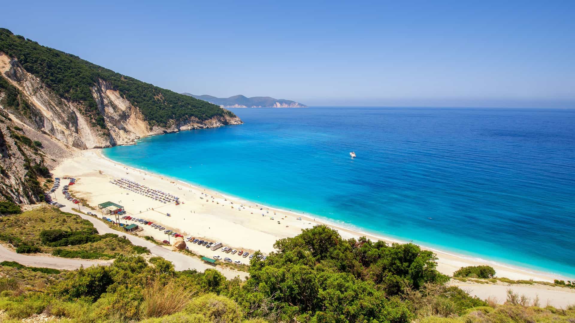 A panoramic view of the stunning Myrtos Beach on Cephalonia, Greece, with its dramatic white pebble coastline, turquoise water, and a steep limestone cliffside.