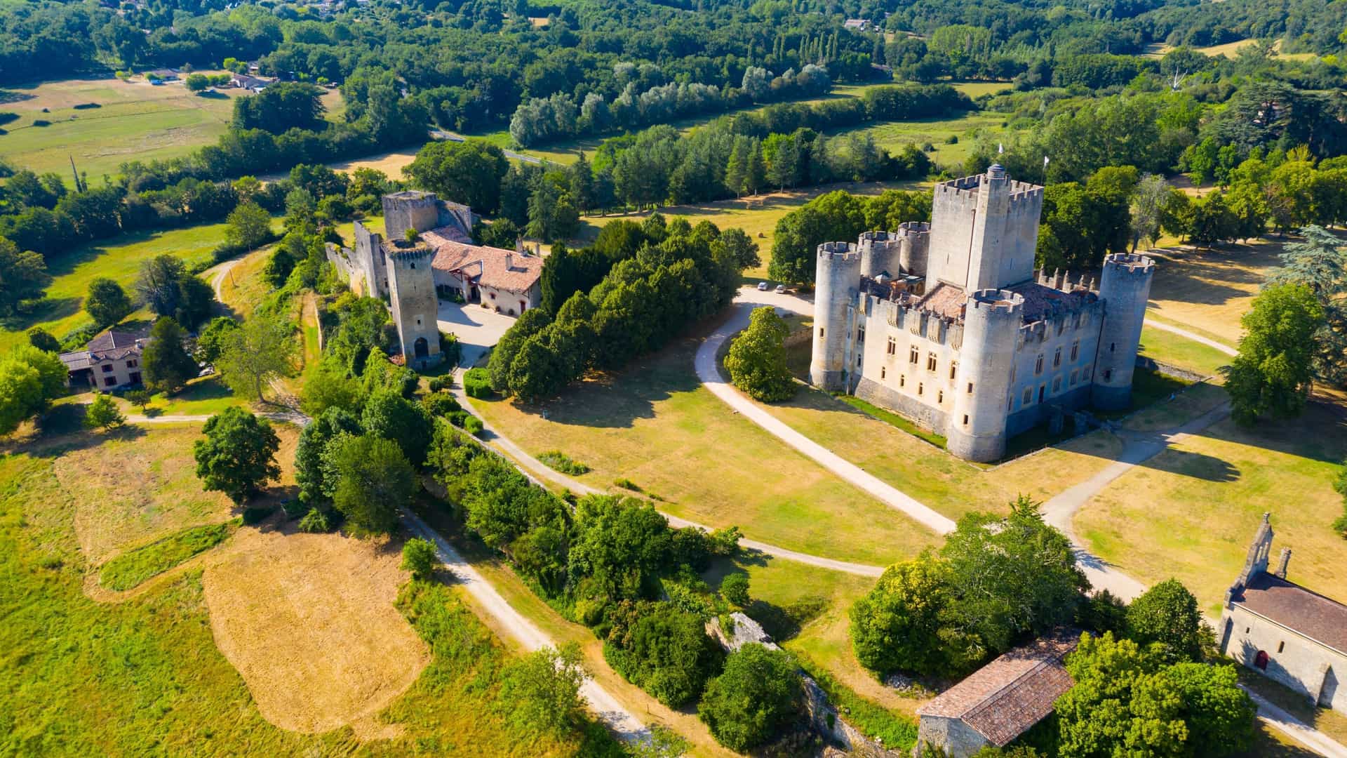 An aerial view of the medieval Château de Roquetaillade, a large castle with multiple towers and courtyards, surrounded by lush green forests and fields in France.