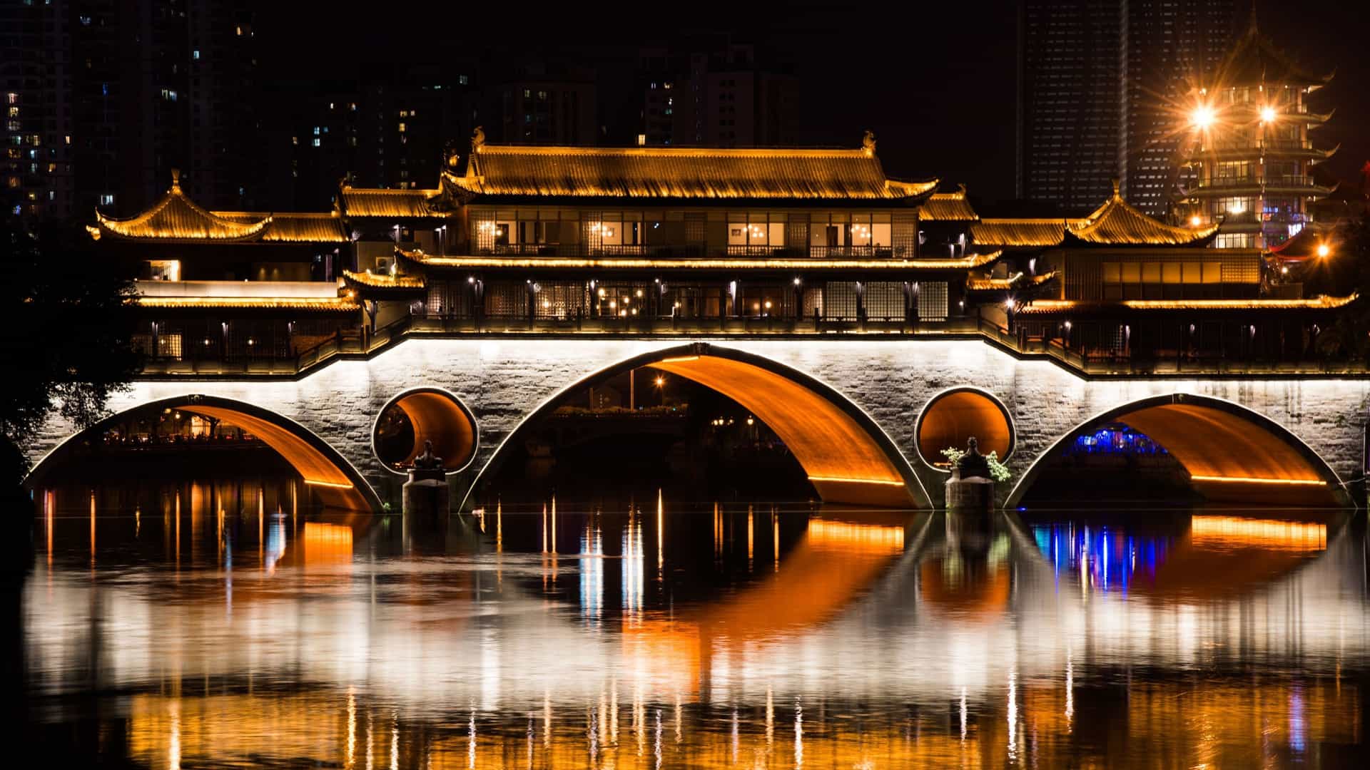 A wide-angle, long exposure shot of the Anshun Bridge in Chengdu, China, brilliantly illuminated at night, with its lights reflecting on the calm river water.