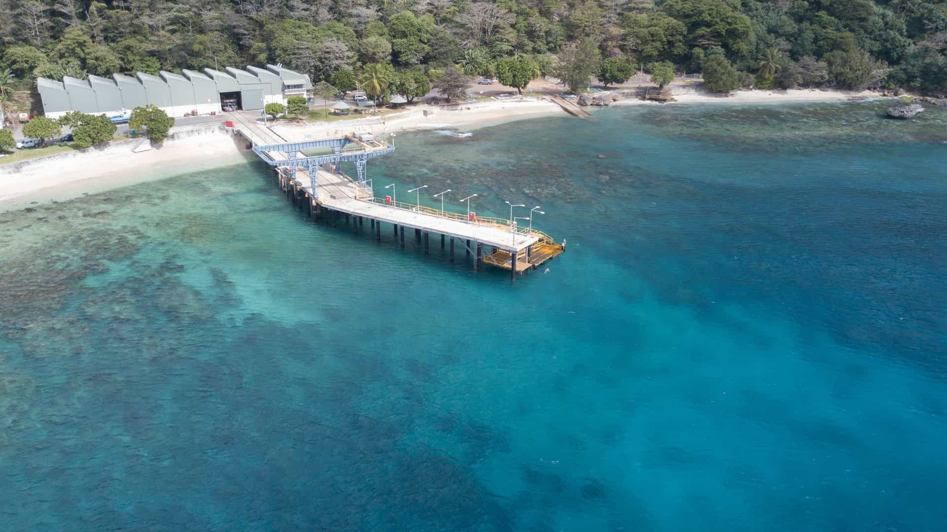 An aerial view of the Christmas Island harbor, showcasing a long concrete pier extending into the brilliant turquoise water, with a sandy beach and lush tropical forest on the shore.