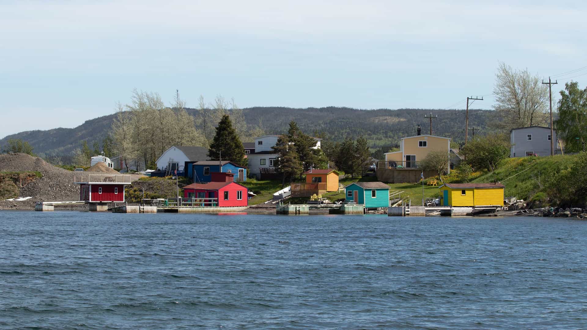 A view from the water of a row of colorful seaside huts and houses in Clarenville, Newfoundland, Canada, with a backdrop of green hills and a clear sky. 
