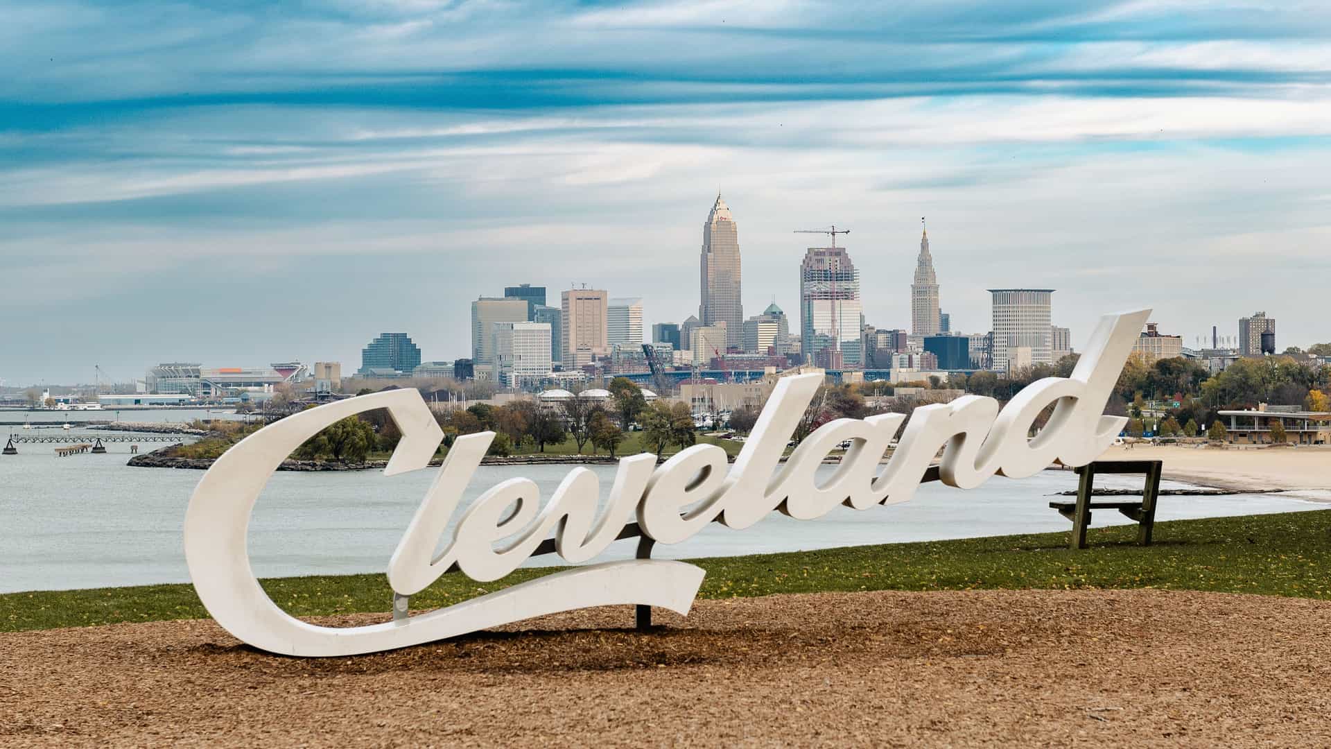A panoramic shot of the Cleveland skyline, with its downtown skyscrapers and Lake Erie in the background, featuring the large white "Cleveland" sign in the foreground.