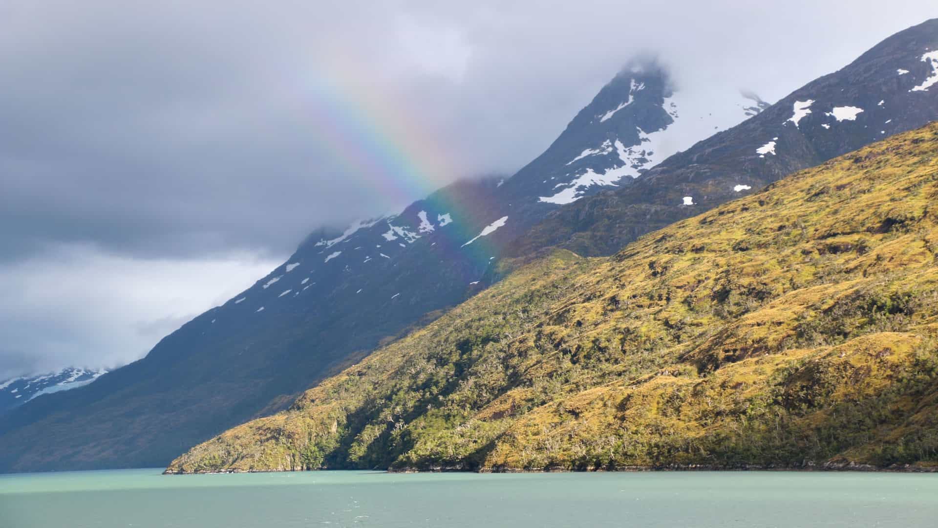 A beautiful landscape of the Cockburn Channel, featuring a rainbow arching over a snow-capped mountain slope, while a grassy hillside descends to the calm turquoise water.