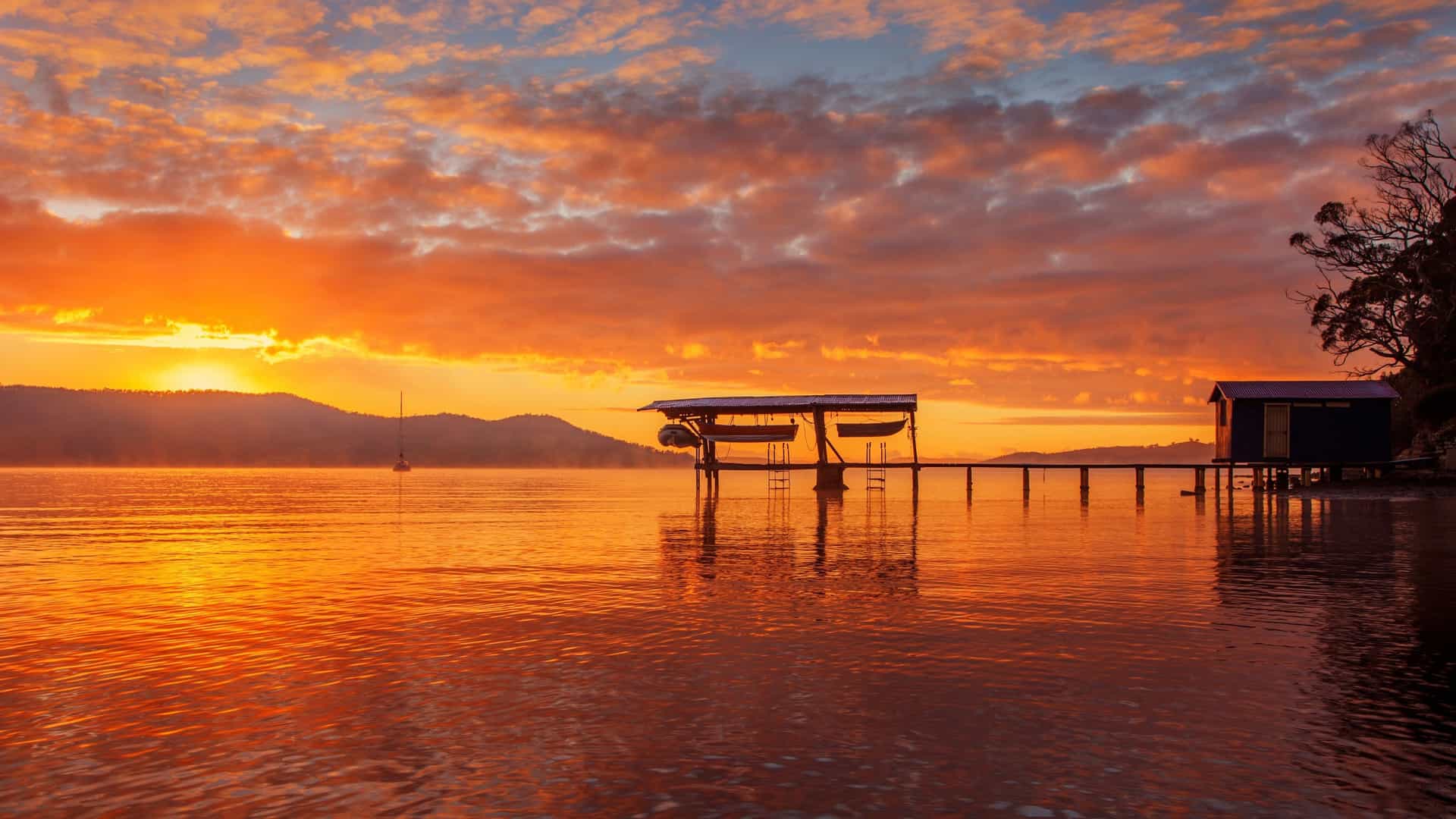 A serene sunrise at Coningham Bay, Tasmania, featuring a wooden jetty with moored boats silhouetted against a fiery orange and red sky over the calm water.