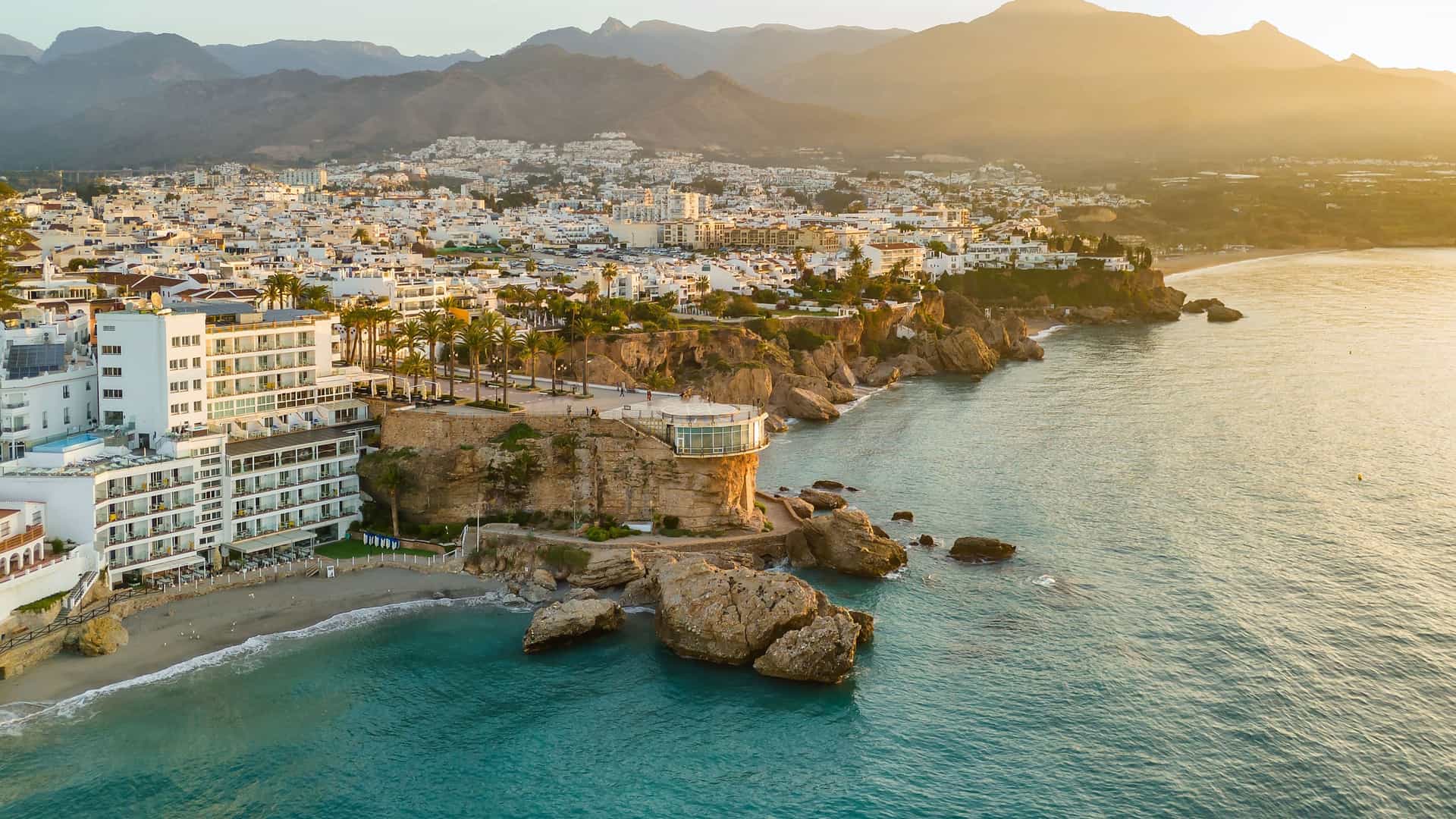 "An aerial view of the coastal town of Nerja on the Costa del Sol in Spain, showcasing white buildings perched on rocky cliffs above a small beach and the turquoise waters of the Mediterranean Sea at sunset. "