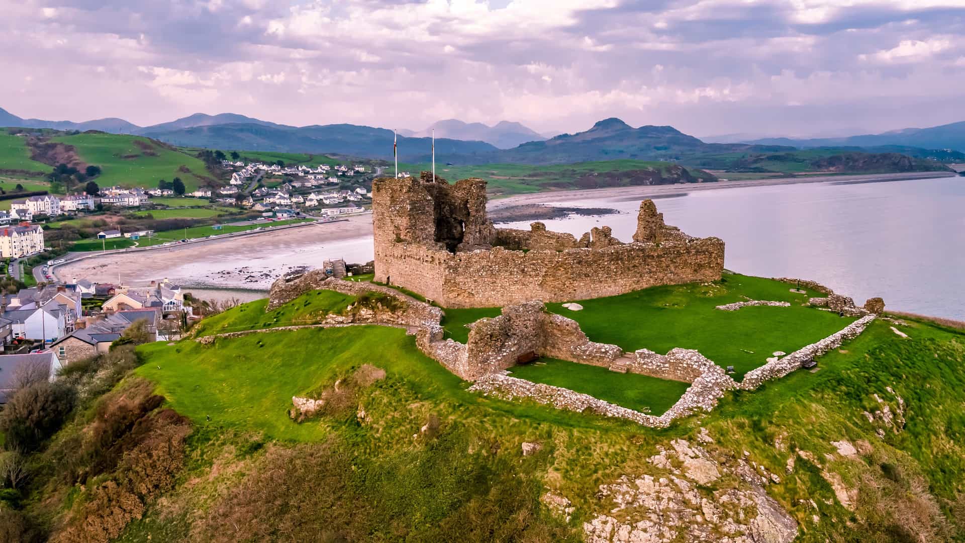 "An aerial view of the historic Criccieth Castle ruins, with its stone walls sitting on a grassy headland, overlooking a sandy beach and the Irish Sea in Wales, with a small town and green hills in the background.  "