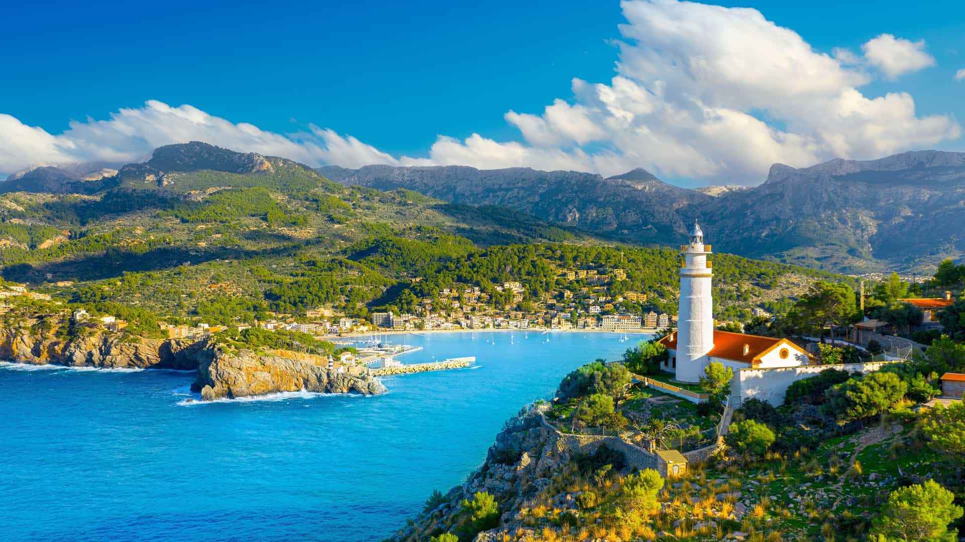A picturesque view of Port de Sóller on Mallorca island, with a white lighthouse on a cliff overlooking the turquoise sea and a tranquil harbor surrounded by mountains.