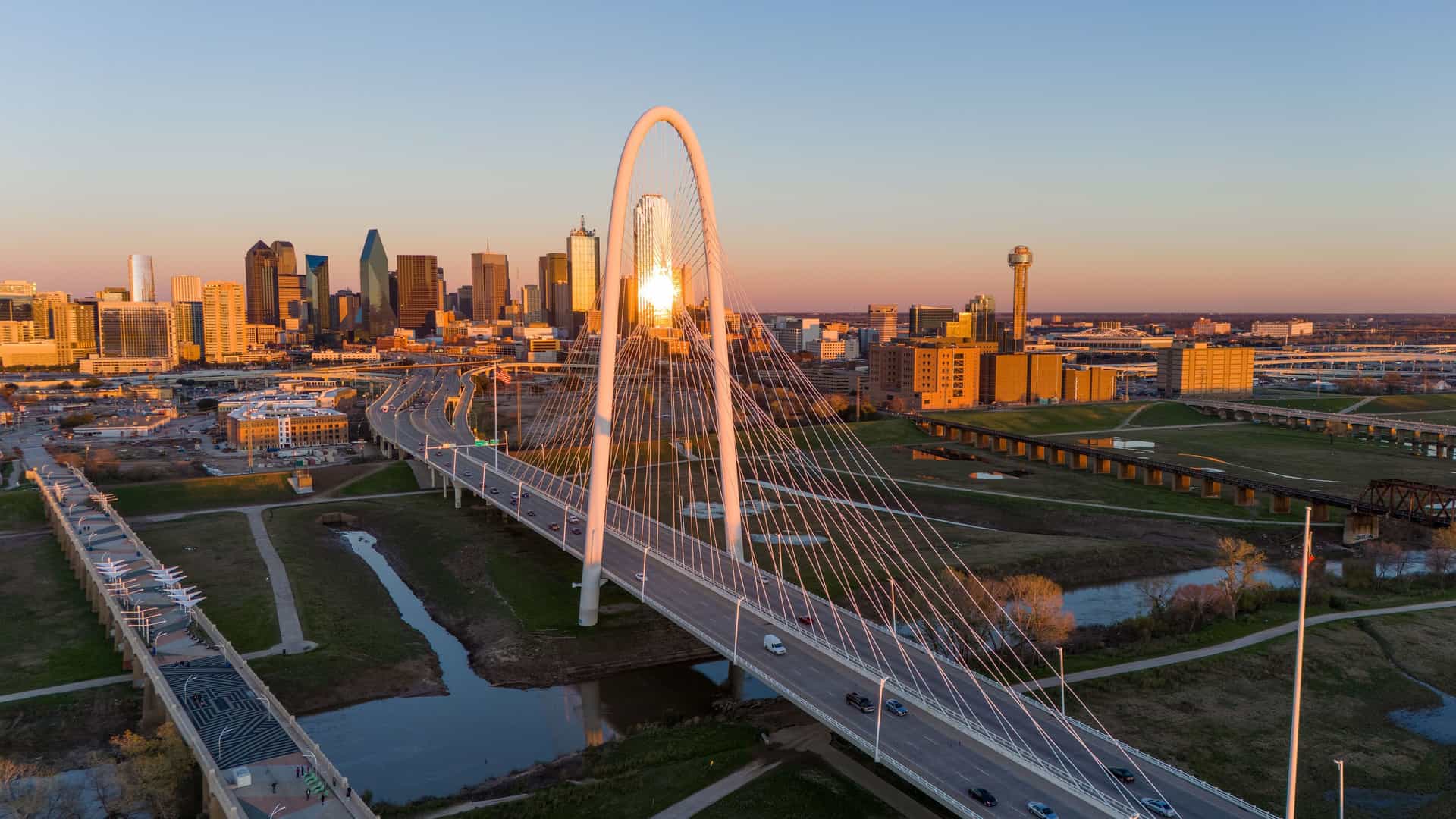 "An aerial shot of the Margaret Hunt Hill Bridge spanning the Trinity River, with cars driving across it at sunset, and the modern skyline of downtown Dallas, Texas, in the background.  "