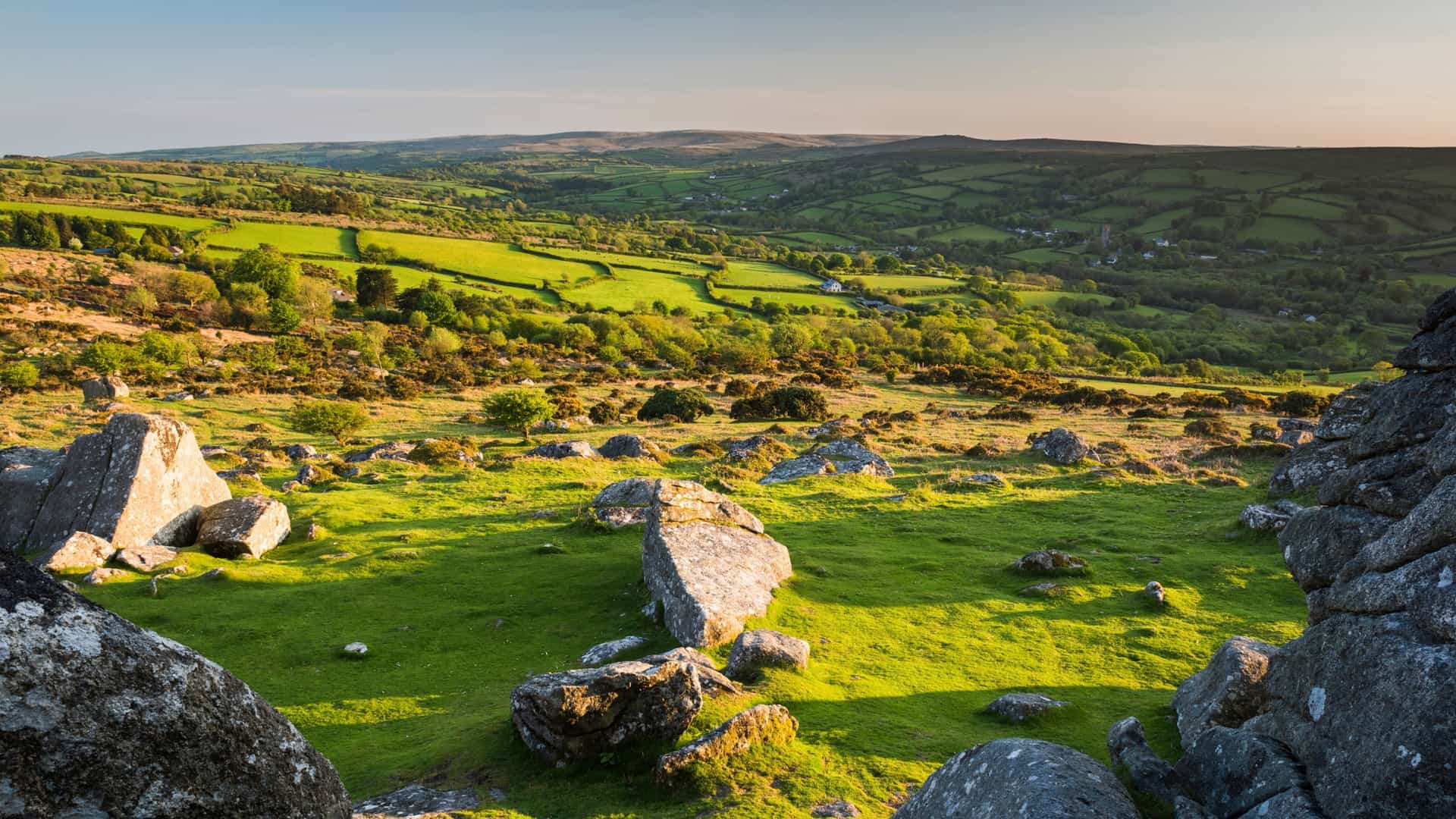 Granite tors and lush green landscape in Dartmoor National Park, Devon, UK. View of farmlands and rolling hills at sunset.