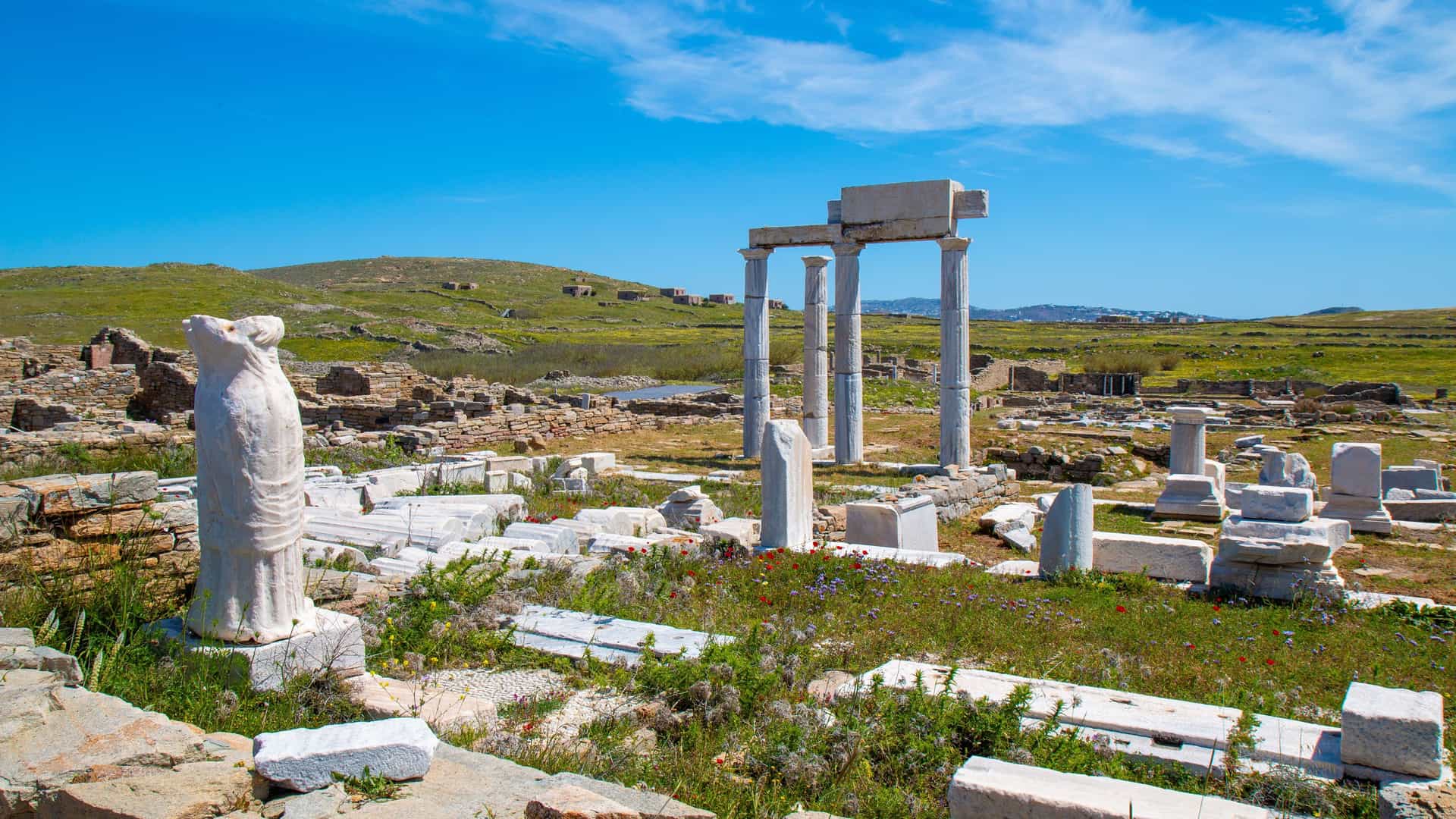 "A scenic shot of the ancient archaeological ruins on the Greek island of Delos, with broken marble columns and statues scattered across a grassy field under a bright blue sky.  "