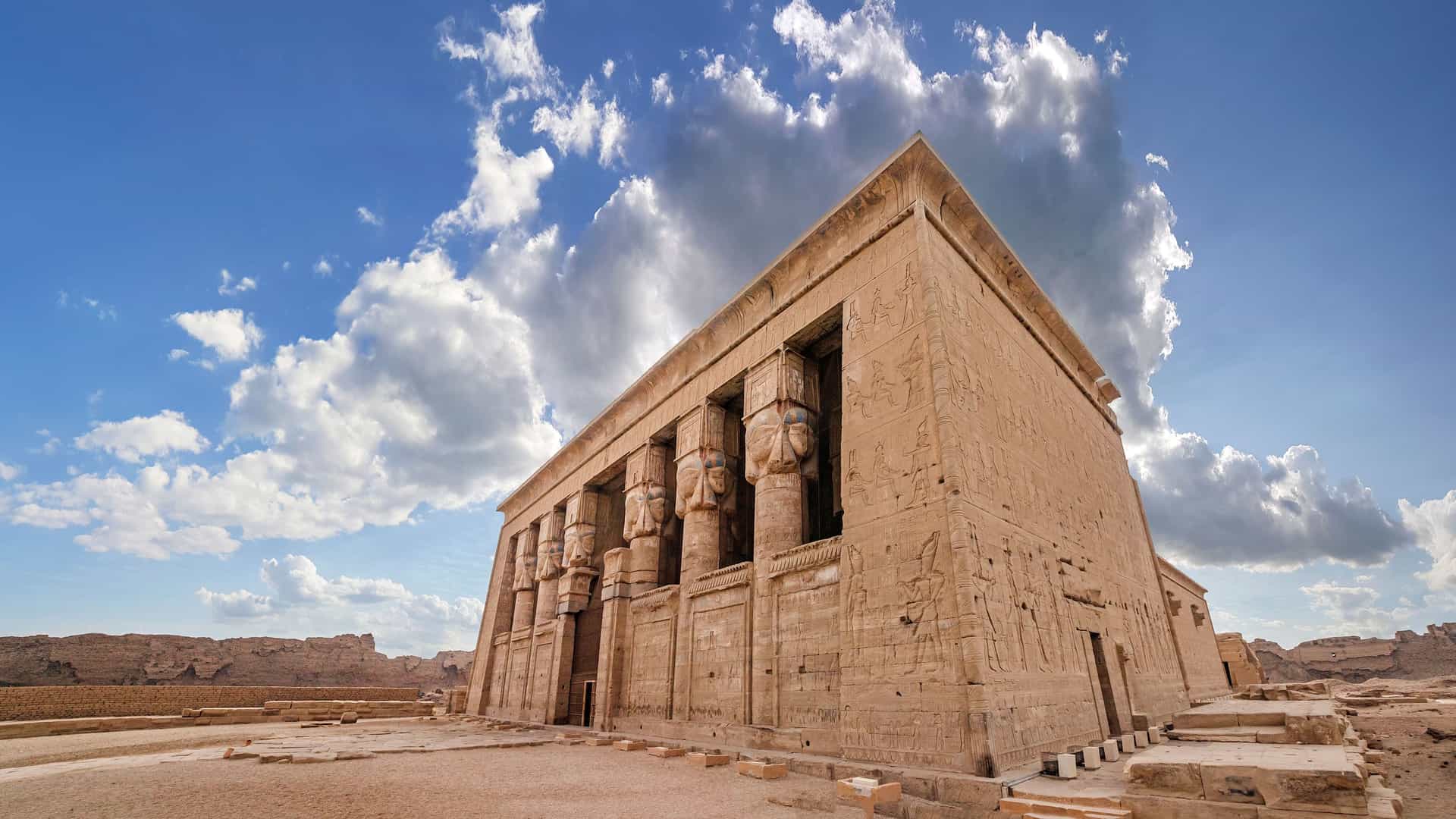 A majestic wide-angle view of the ancient Temple of Hathor in Dendera, Egypt, featuring carved columns and intricate hieroglyphics on its walls under a dramatic blue and cloudy sky. 