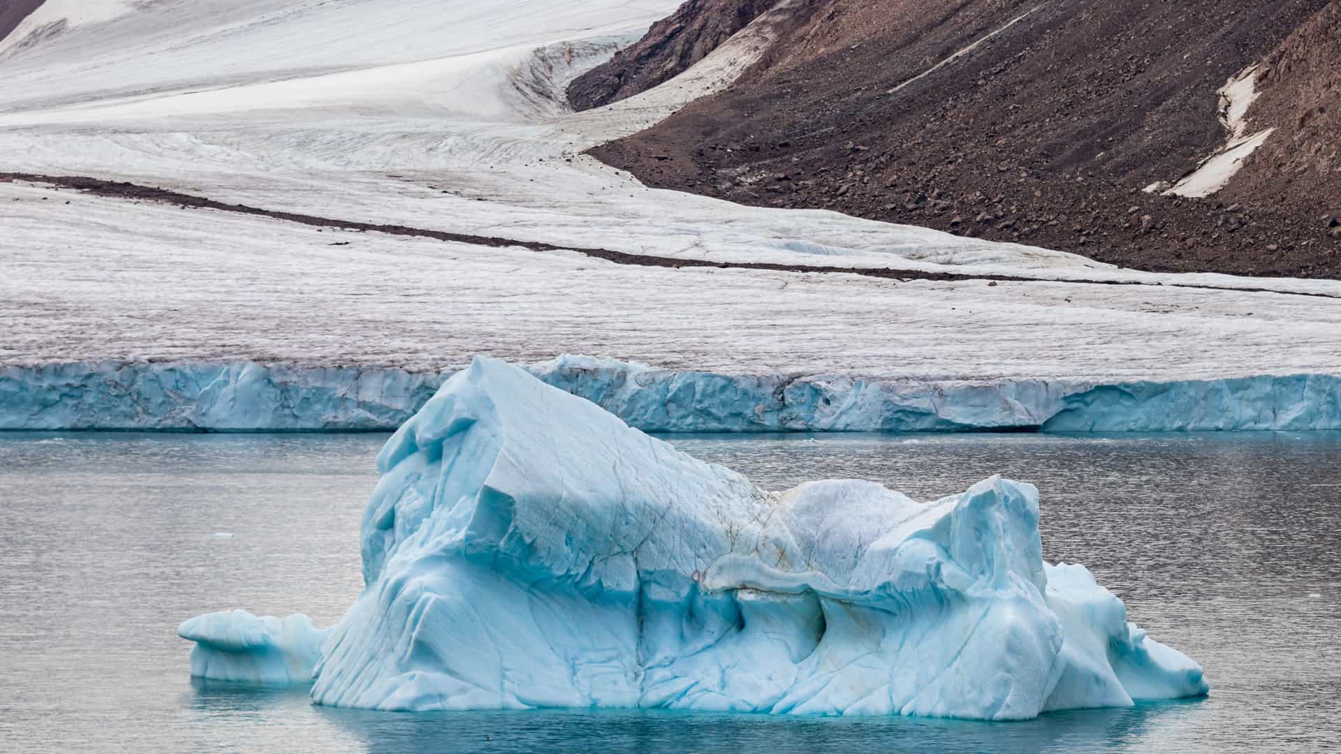 "A large, blue iceberg floating in the frigid waters of the Arctic Ocean, with a massive glacier and a dark brown mountain in the background on Diana Island, Nunavut, Canada.  "