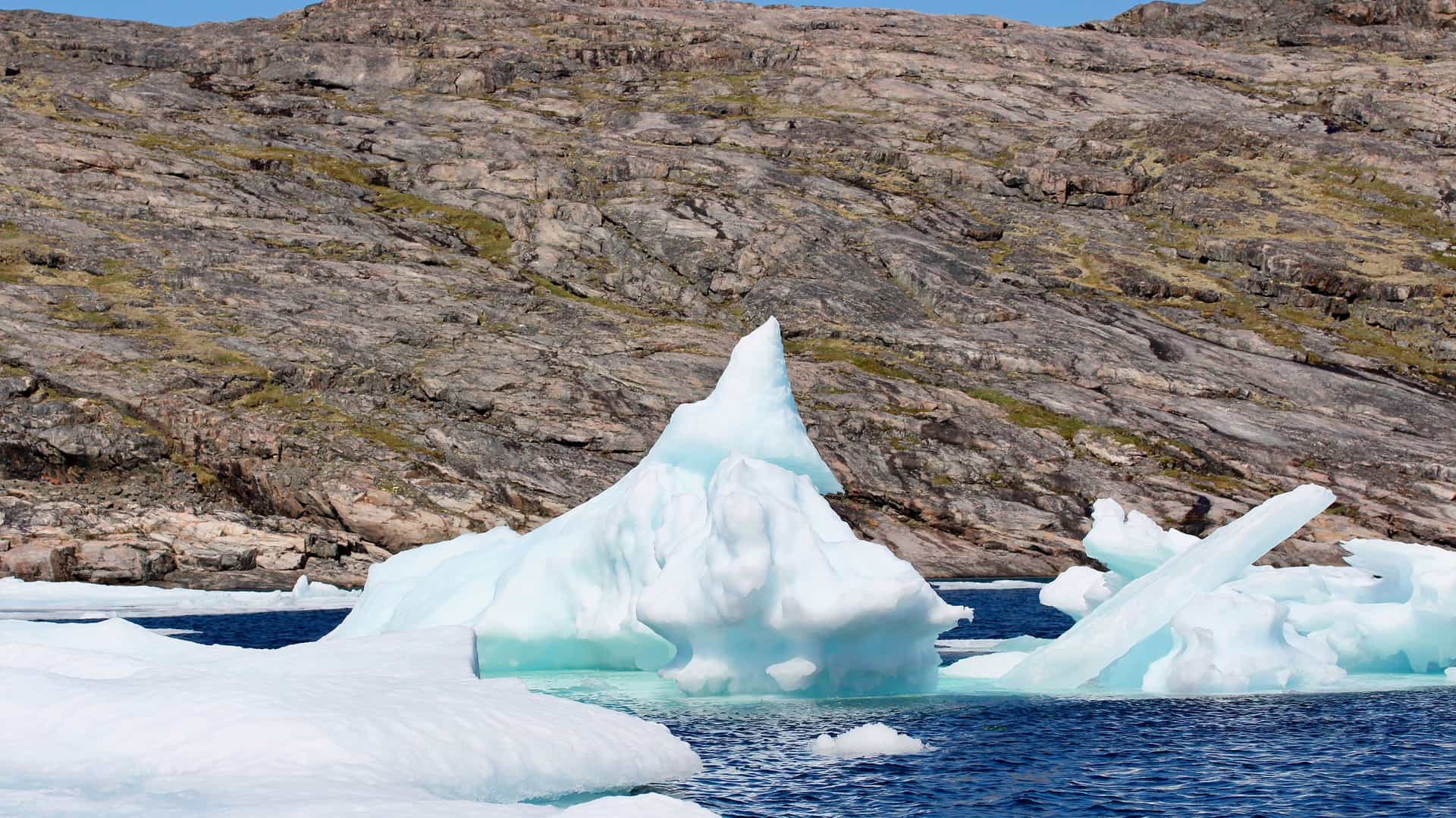 A massive, pointed iceberg with a pale blue hue floats in the deep blue water of the Arctic, set against the barren, rocky landscape of Digges Island, Nunavut, Canada.