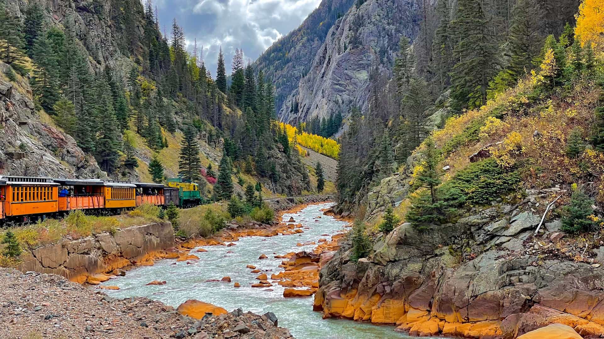 "A vibrant scene of the historic Durango & Silverton Narrow Gauge Railroad train traveling along a track beside the Animas River in a narrow canyon. The surrounding slopes are covered in pine and golden aspen trees in the autumn.  "