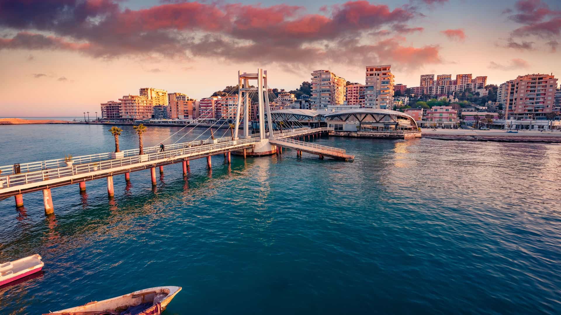"A vibrant scenic view of the coastal city of Durrës, Albania, with a modern, white pedestrian bridge crossing the harbor toward the city's waterfront buildings at sunset.  "