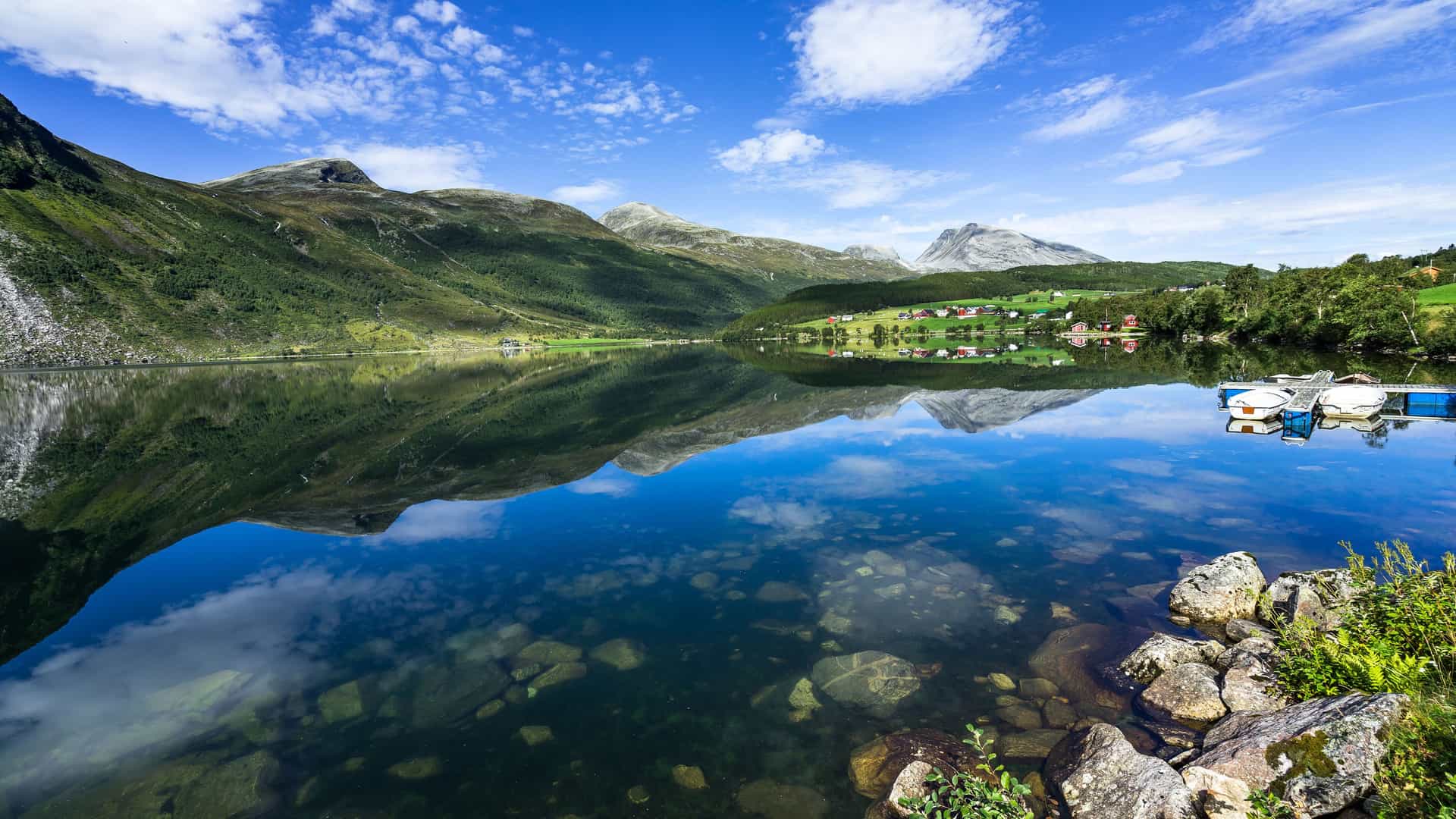 "A scenic view of the tranquil lake and towering mountains of Eidsdal, Norway, with scattered houses and a small dock with boats on the far shore, all perfectly reflected in the clear, still water under a beautiful blue sky.  "