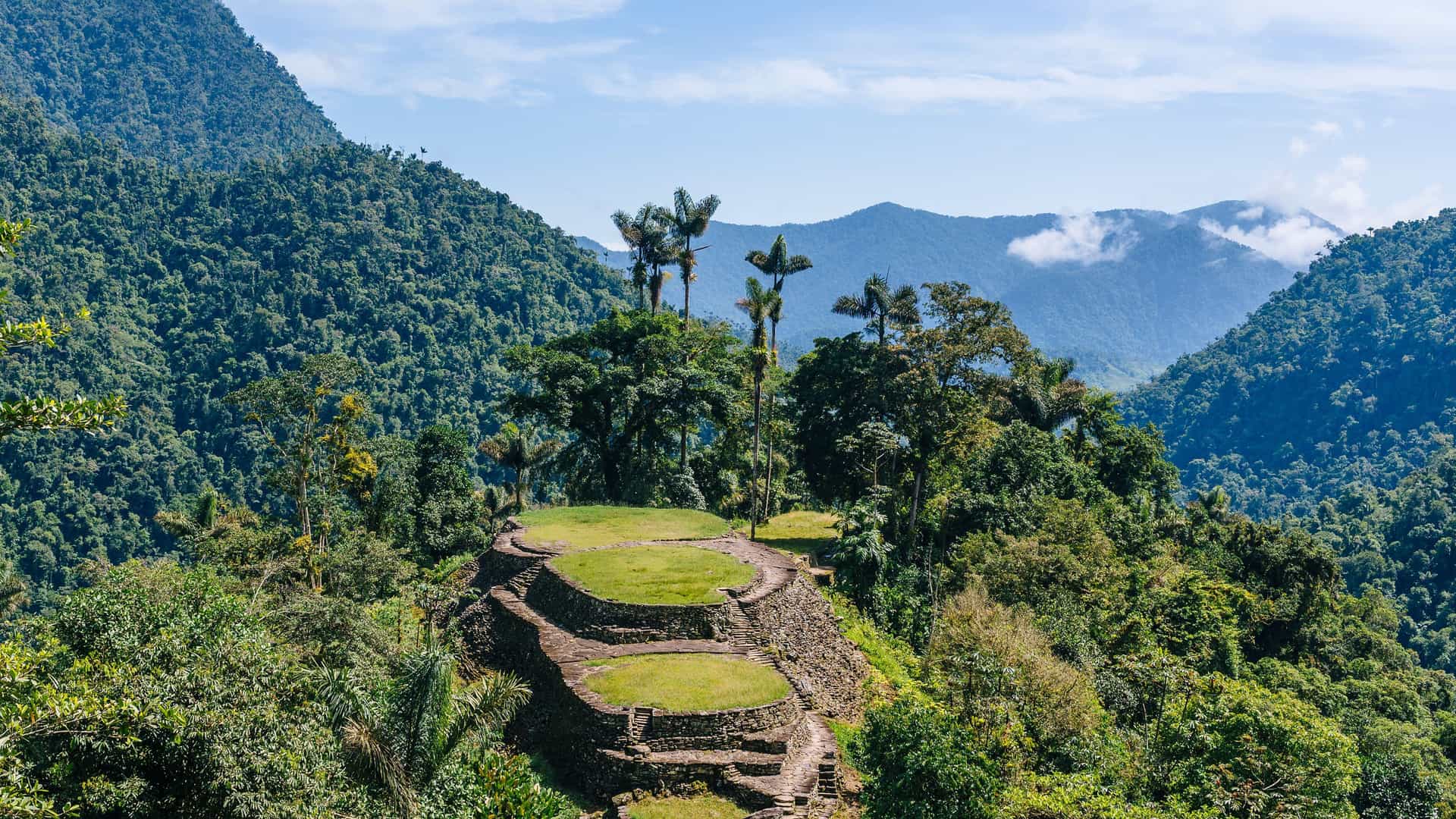 A stunning aerial shot of the Ciudad Perdida ruins near El Banco, Colombia, with ancient stone terraces and palm trees nestled in the lush, green mountains.