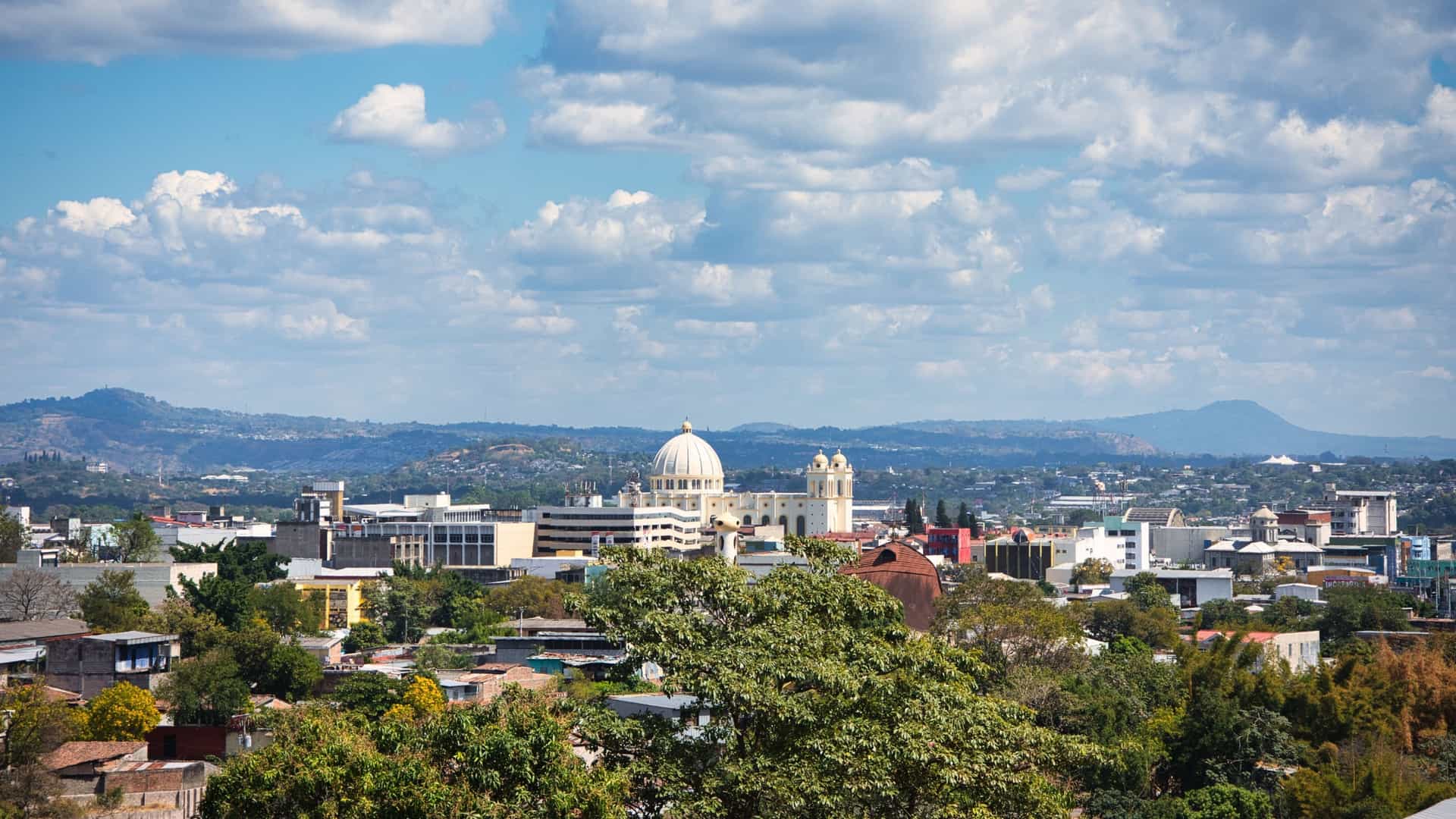 A scenic overview of San Salvador City, the capital of El Salvador, captured at sunset or dusk. The cityscape of buildings and infrastructure fills the valley floor and slopes, extending towards the backdrop. The sky displays vibrant sunset colors, and in the distance, a prominent volcano, likely the San Salvador Volcano (or El Boquerón), rises above the urban area, emphasizing the city's geographical location in Central America.