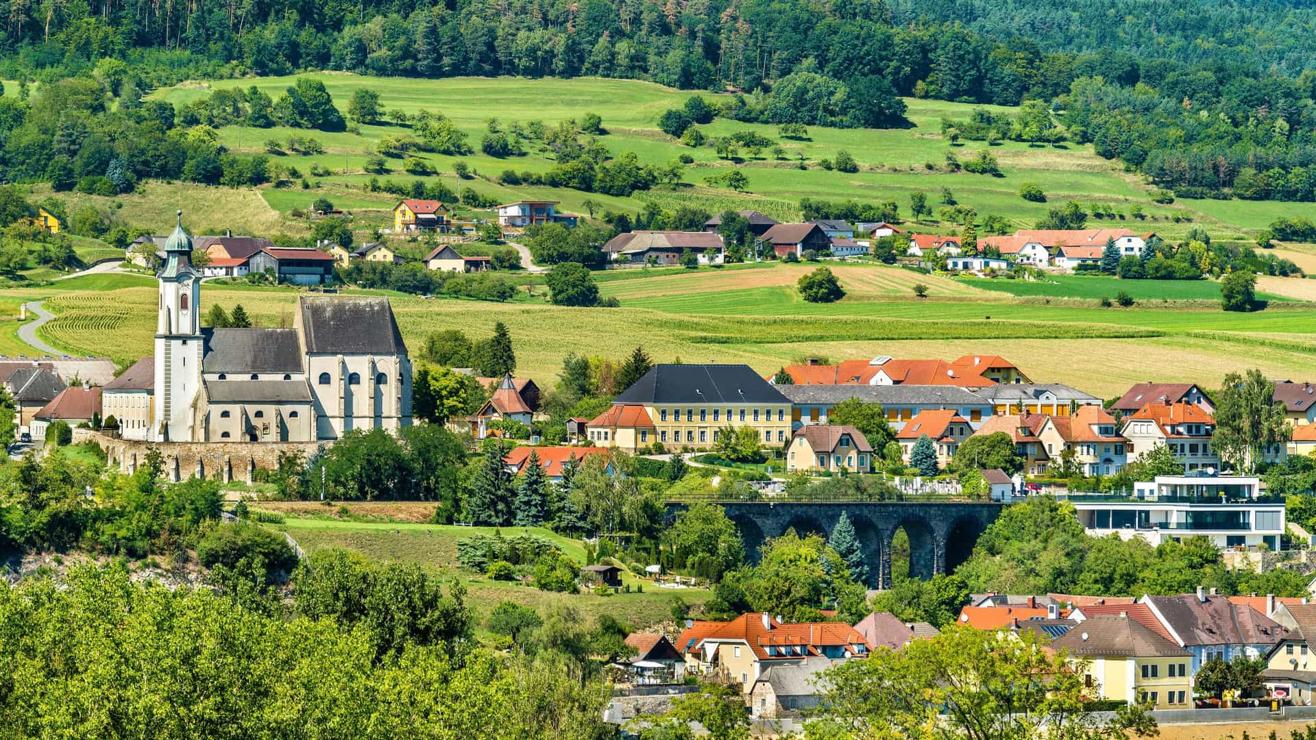 A beautiful pastoral scene of the village of Emmersdorf in Austria, with a historic stone church and other buildings nestled in a lush green valley with rolling hills.