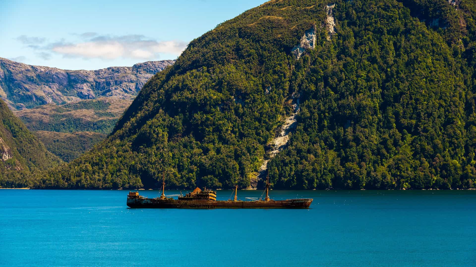 A rusting shipwreck sits in the calm turquoise waters of English Narrows, Chile, with towering, tree-covered mountains and a bright blue sky providing a beautiful backdrop.