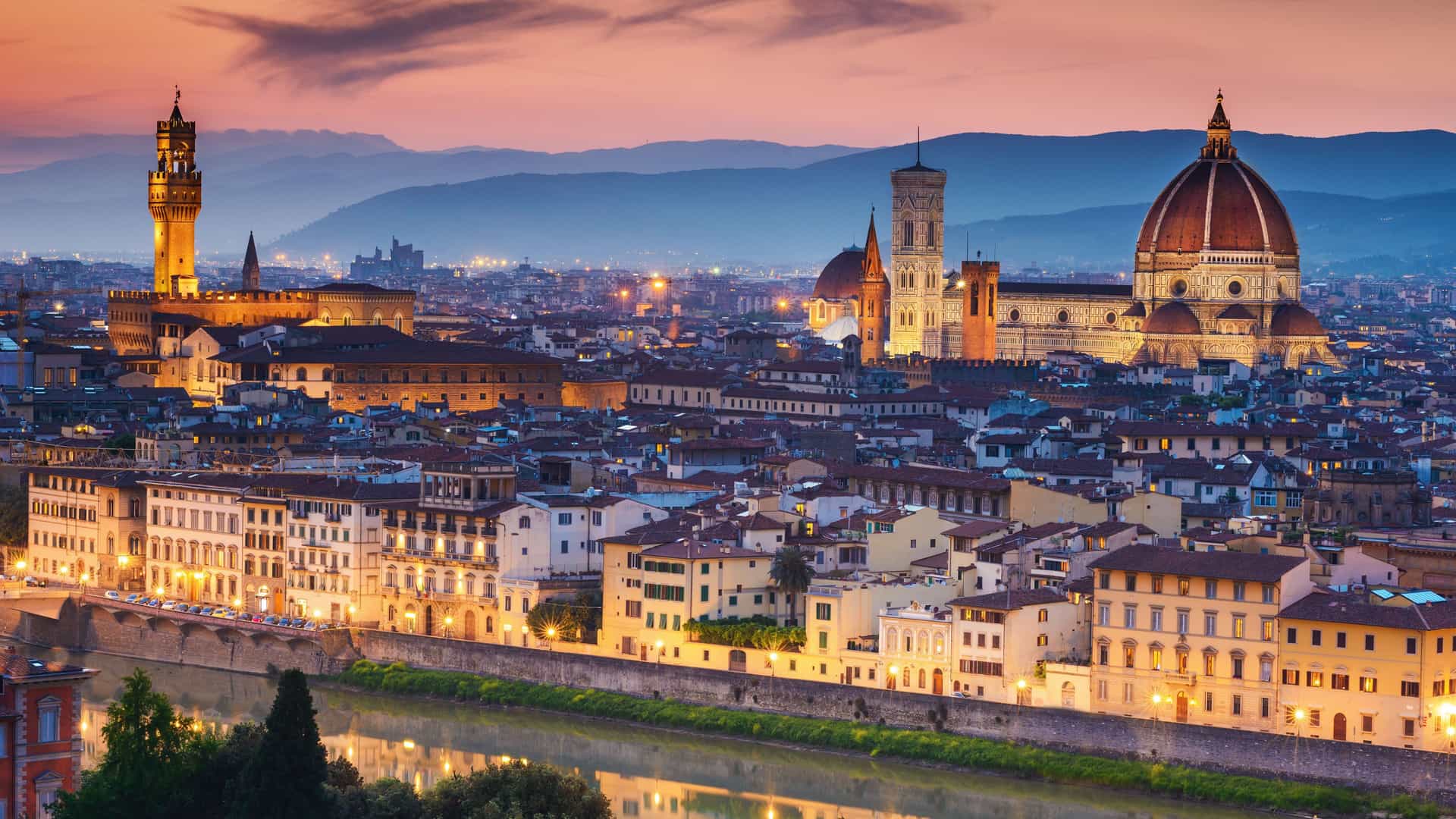 A beautiful twilight cityscape of Florence, Italy, with the iconic Duomo and Ponte Vecchio visible along the Arno River, and the cityscape's warm lights illuminating the historic buildings as the sky turns a vibrant orange.