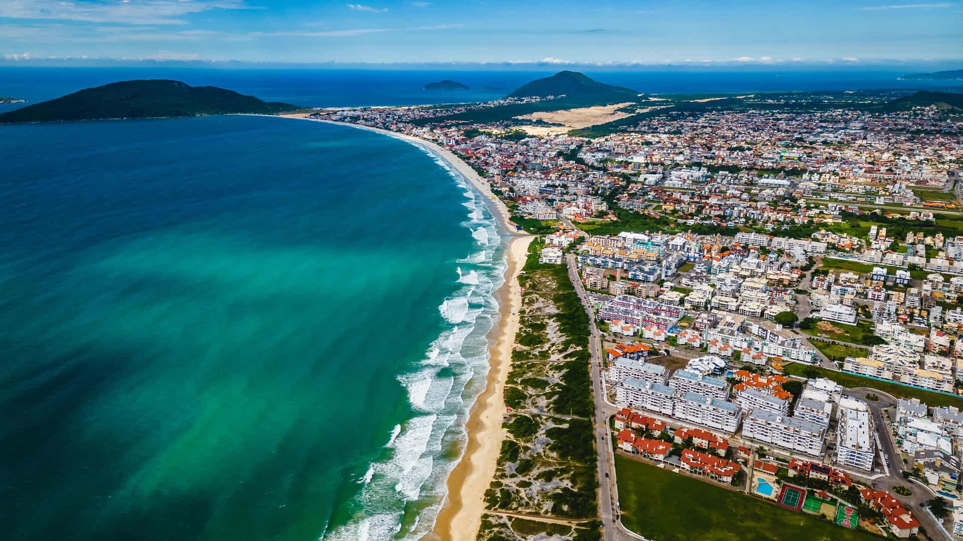 An aerial view of Florianópolis, Brazil, showing a long, wide sandy beach with foamy waves, a bustling coastal city, and lush green hills stretching out along the shoreline.