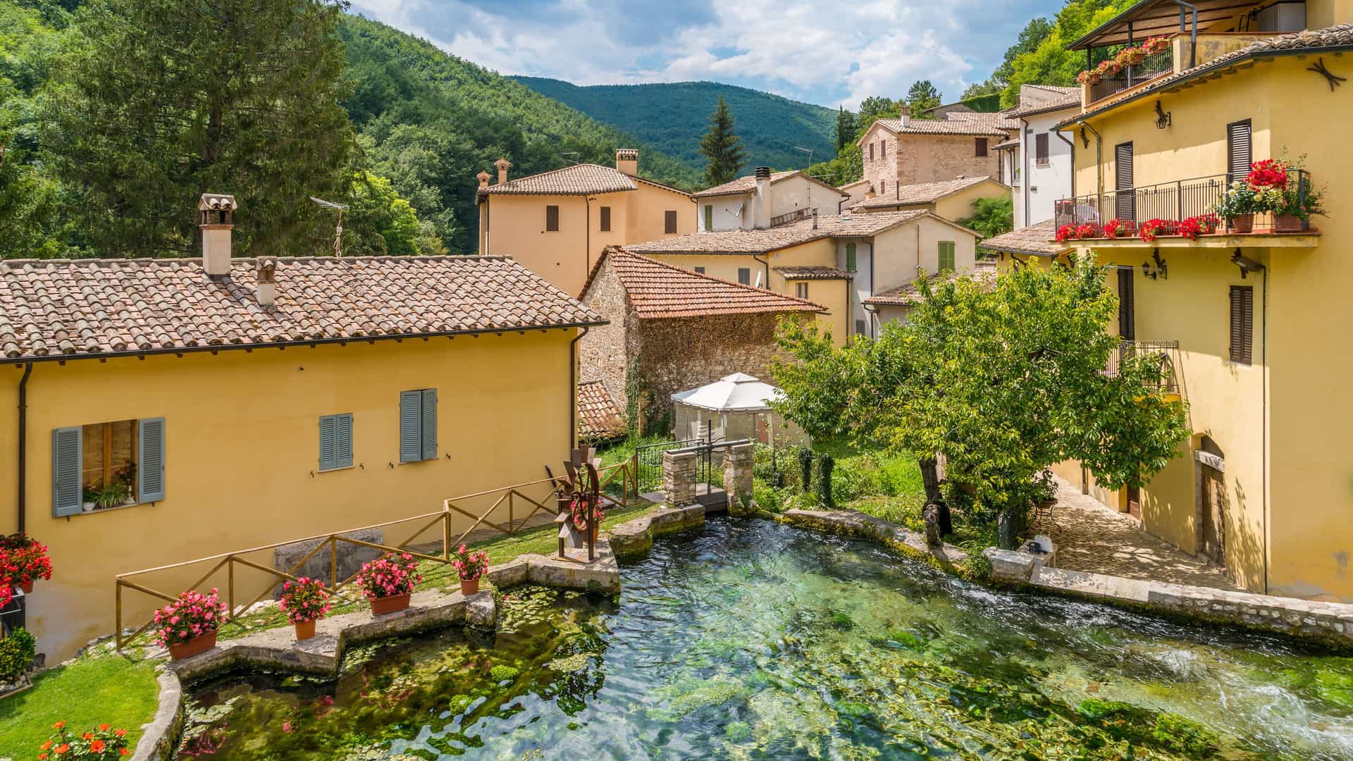 "A tranquil view of Rasiglia, a small village near Foligno, Italy, featuring a crystal-clear stream running through the center of historic stone and stucco houses on a sunny day.  "