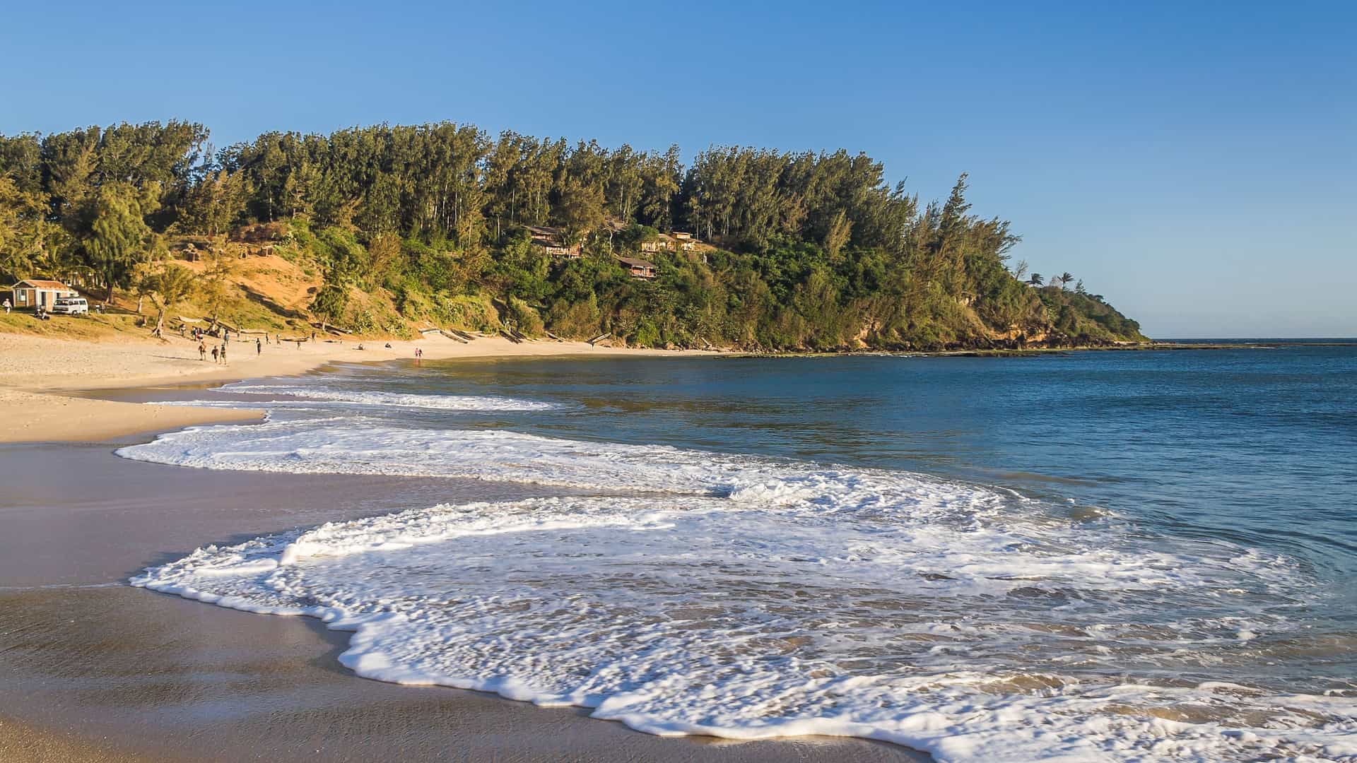 A tranquil beach with gentle waves on the coast of Fort Dauphin, also known as Tôlanaro, Madagascar, with a lush forest and a few people enjoying the sunny afternoon.