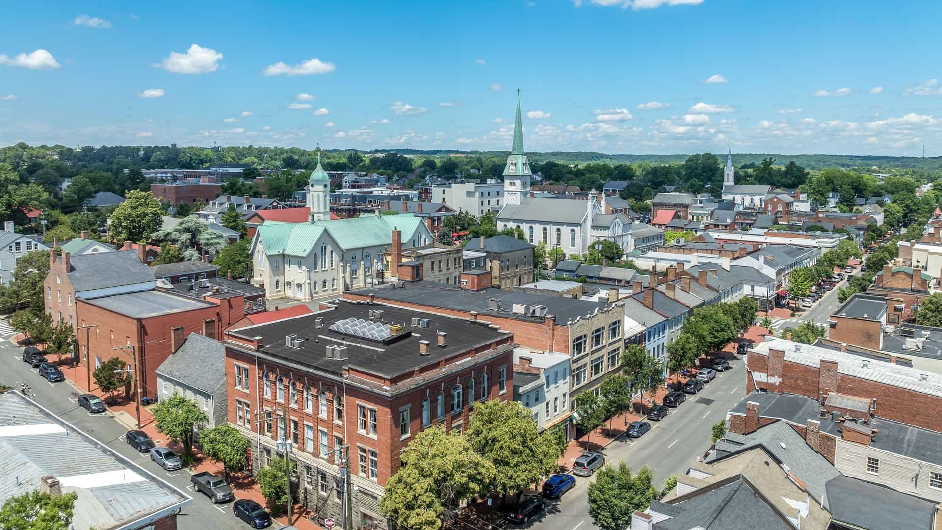 "An aerial view of the historic downtown area of Fredericksburg, Virginia, with its blend of red brick and colonial-style buildings and prominent church steeples under a bright blue sky.  "