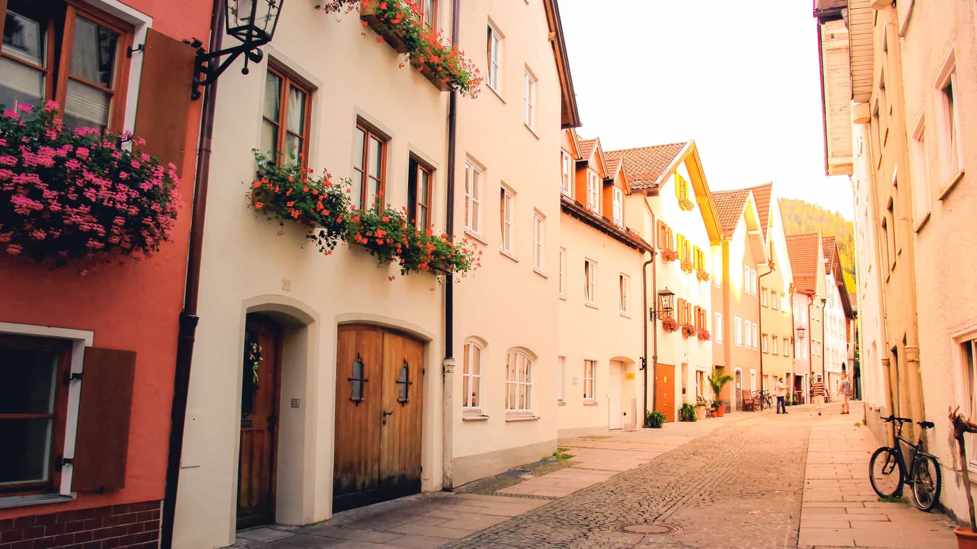"A narrow, cobbled street in the charming town of Füssen, Germany, with a line of traditional buildings with bright window boxes full of red flowers and a person with a bicycle.  "
