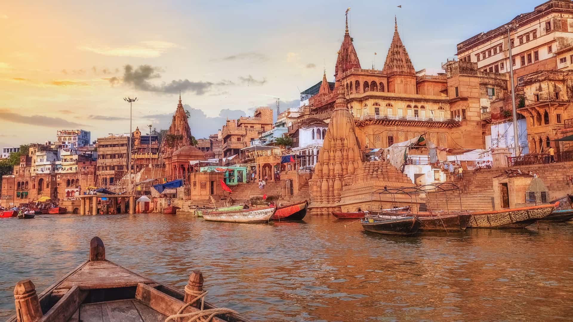 "A wide-angle view of the Ganges River in Varanasi, India, with a row of ancient buildings, temples, and ghats descending to the water's edge, all under a warm evening sky.  "