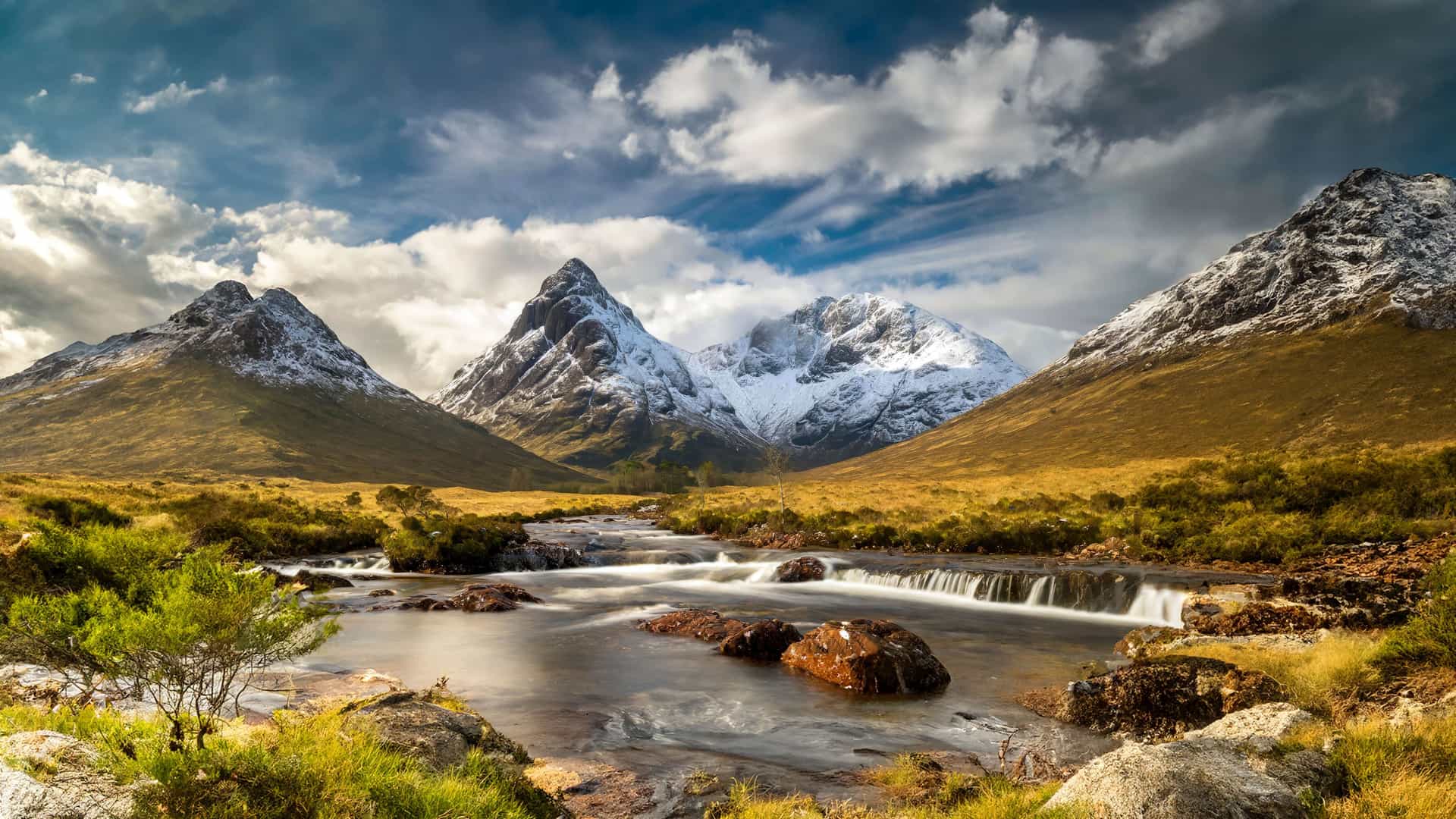"A dramatic landscape shot of the Glencoe Valley in the Scottish Highlands, featuring snow-capped mountains and a stream rushing over rocks in the foreground under a dynamic, cloudy sky.  "
