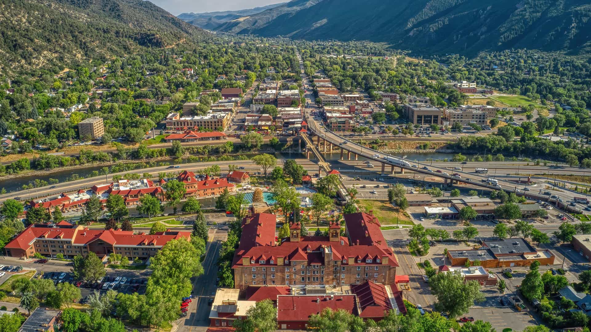 "An aerial view of Glenwood Springs, Colorado, nestled between two mountains, with the prominent Hotel Colorado in the foreground, a highway bridge, and a river flowing through the town.  "