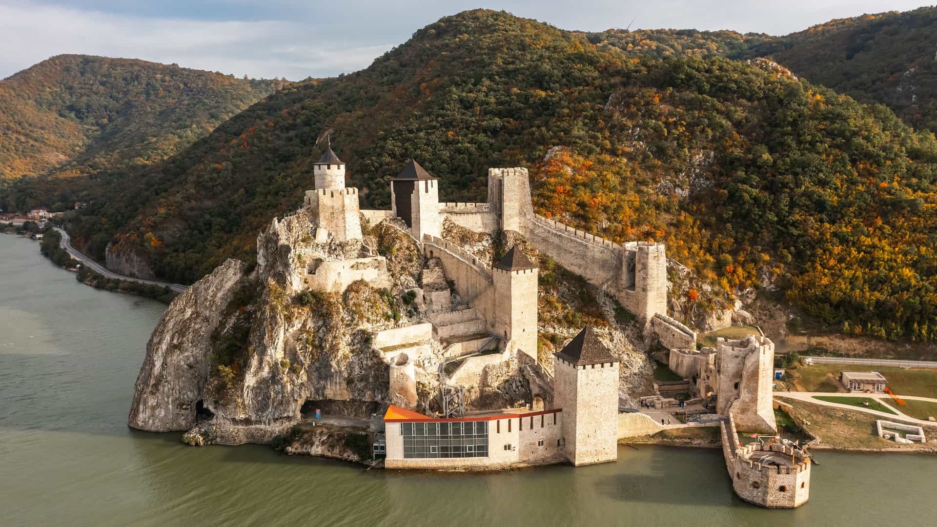 An aerial view of the majestic Golubac Fortress, a medieval castle built on a rocky outcrop along the Danube River, with lush, forested hills in the background.