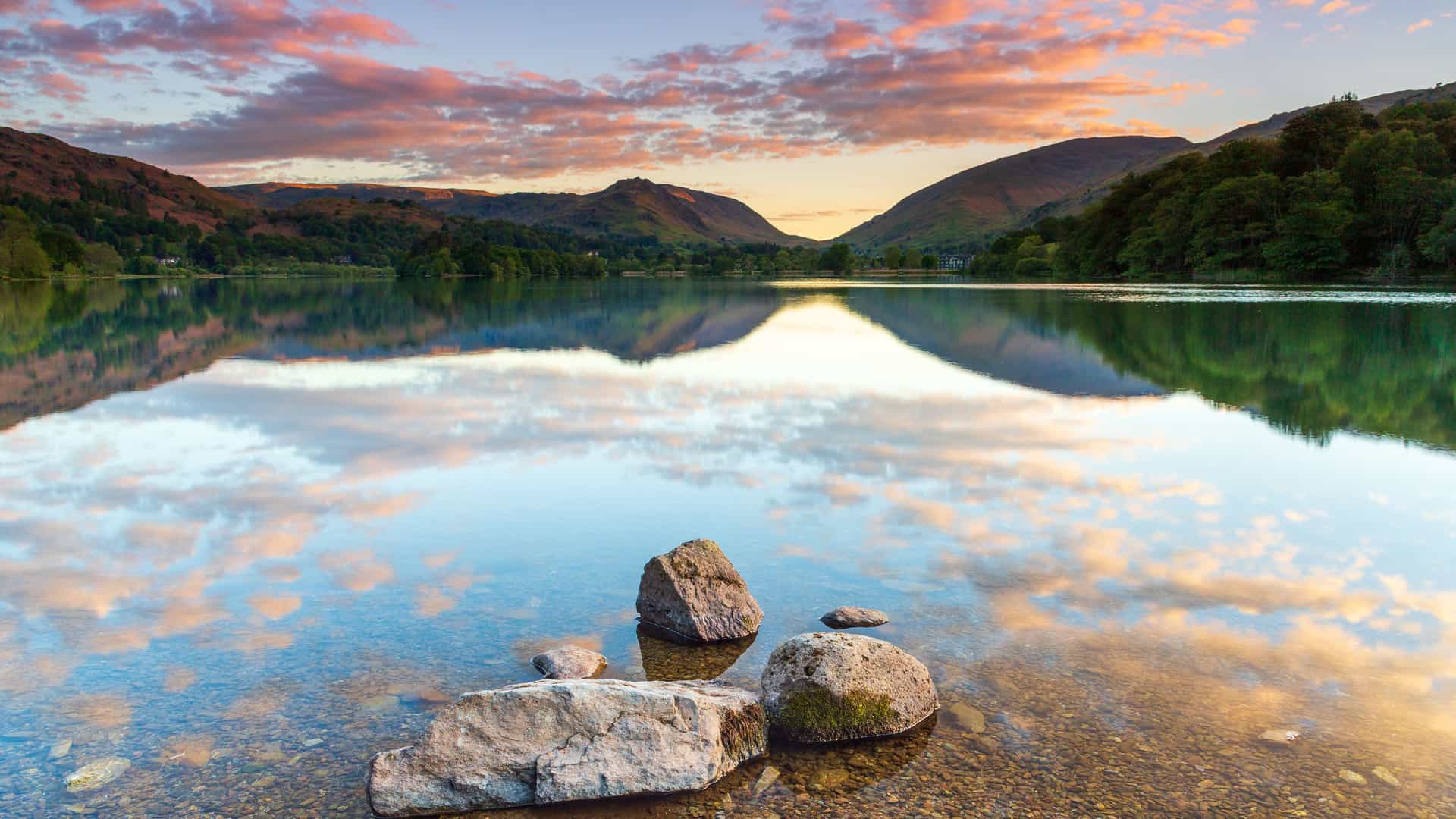 A calm and reflective image of Grasmere Lake in the Lake District, England, at sunset with mountains and a beautiful sky reflecting on the water.