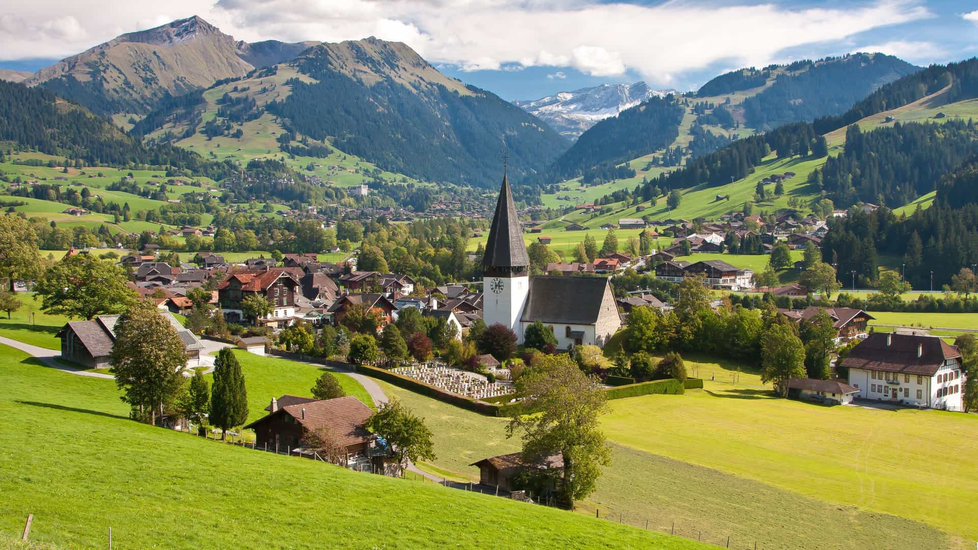 A picturesque view of the Swiss village of Gstaad, with its charming church and traditional chalets nestled in a lush green alpine valley surrounded by mountains.
