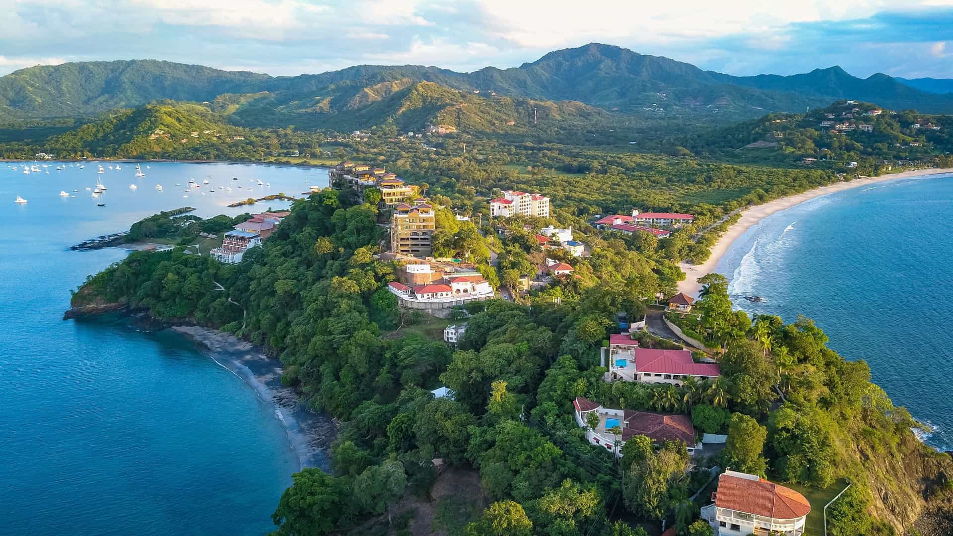"An aerial view of the coastline of Guanacaste, Costa Rica, showing a verdant peninsula with luxury resorts and homes, with a white sand beach and boats moored in the clear blue water.  "