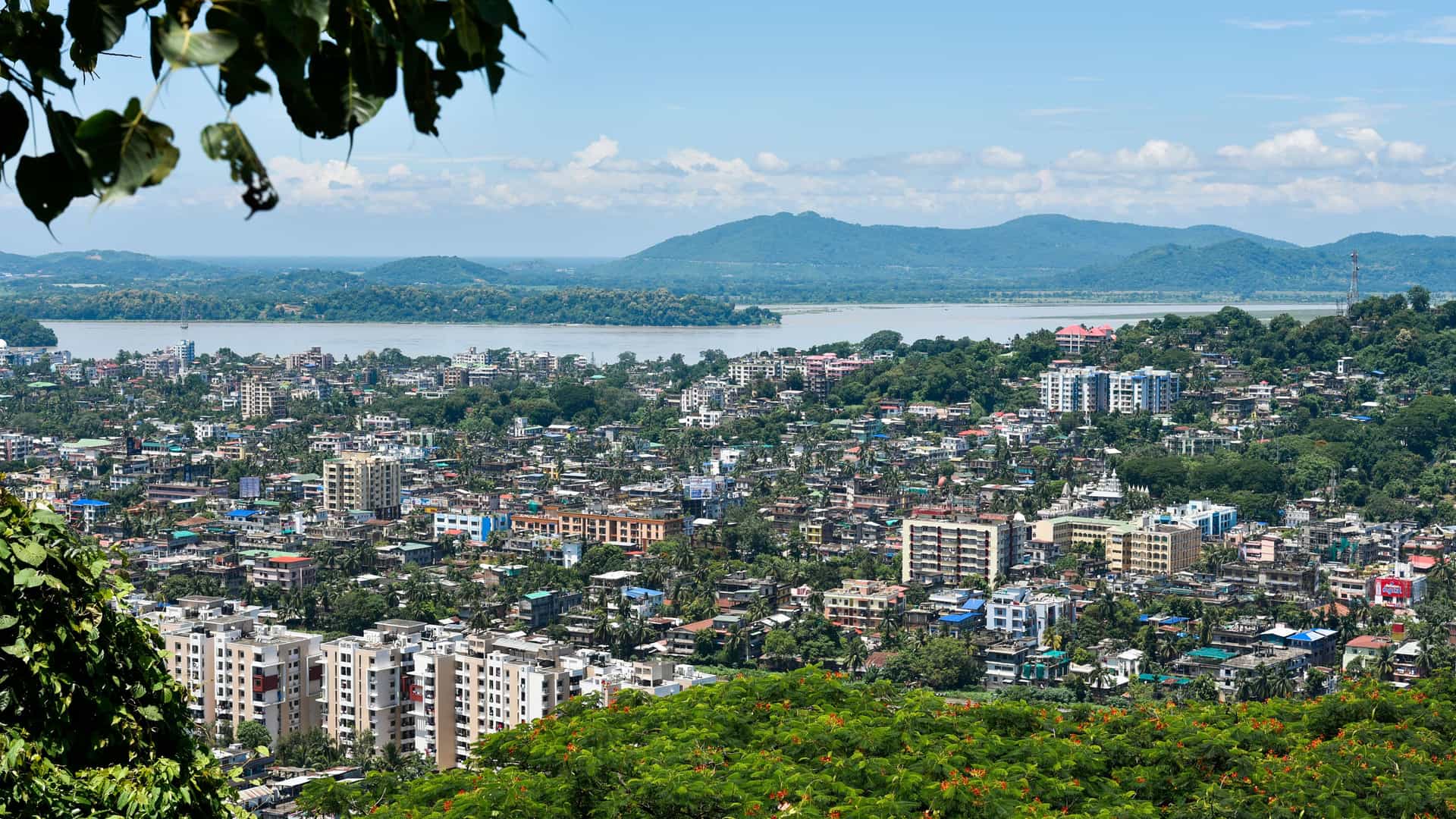 "A scenic, elevated view of Guwahati, India, with the city's buildings nestled among lush trees, the Brahmaputra River in the background, and distant hills under a blue sky.  "