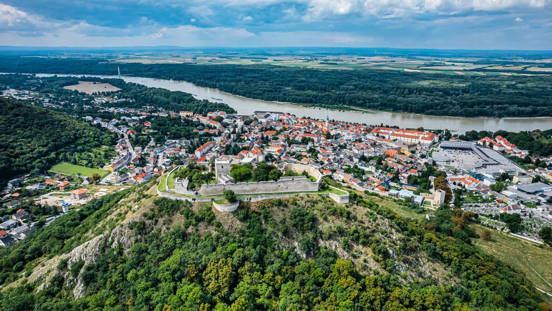 "An aerial view of Hainburg, Austria, with the ruins of the historic Hainburg Castle perched on a hill overlooking the town and the vast Danube River winding through the green landscape.  "