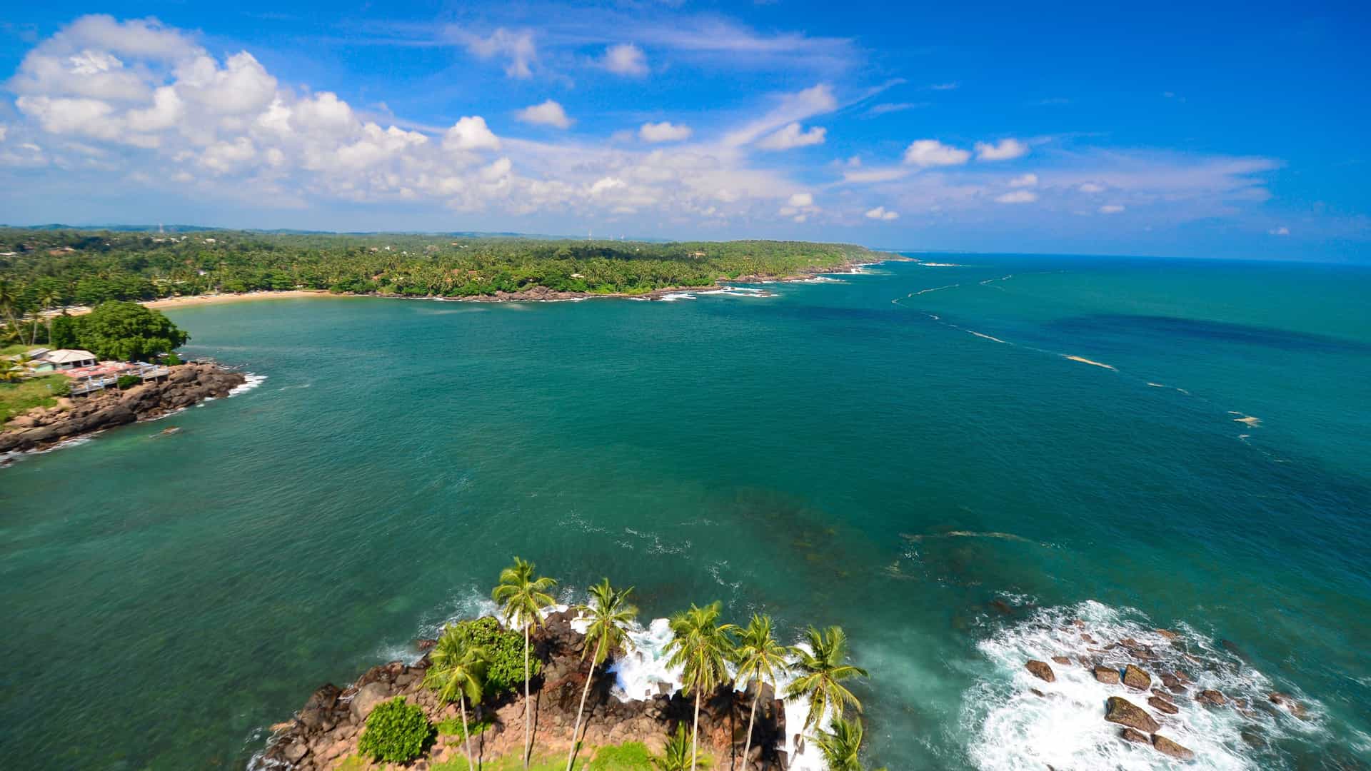 An aerial view of the tropical coastline of Hambantota, Sri Lanka, with lush palm trees on rocky outcrops, a sandy beach, and the expansive turquoise Indian Ocean.