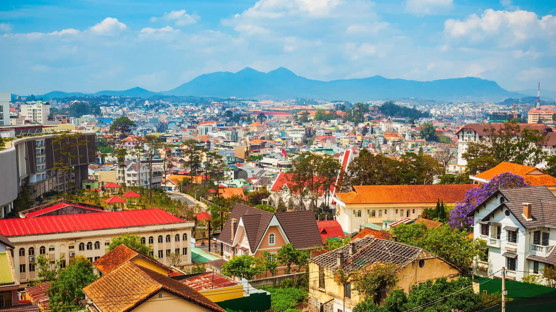 A scenic, elevated view of the city of Da Lat in Vietnam, with a variety of buildings and red-tiled roofs in the foreground and a mountain range in the distance.
