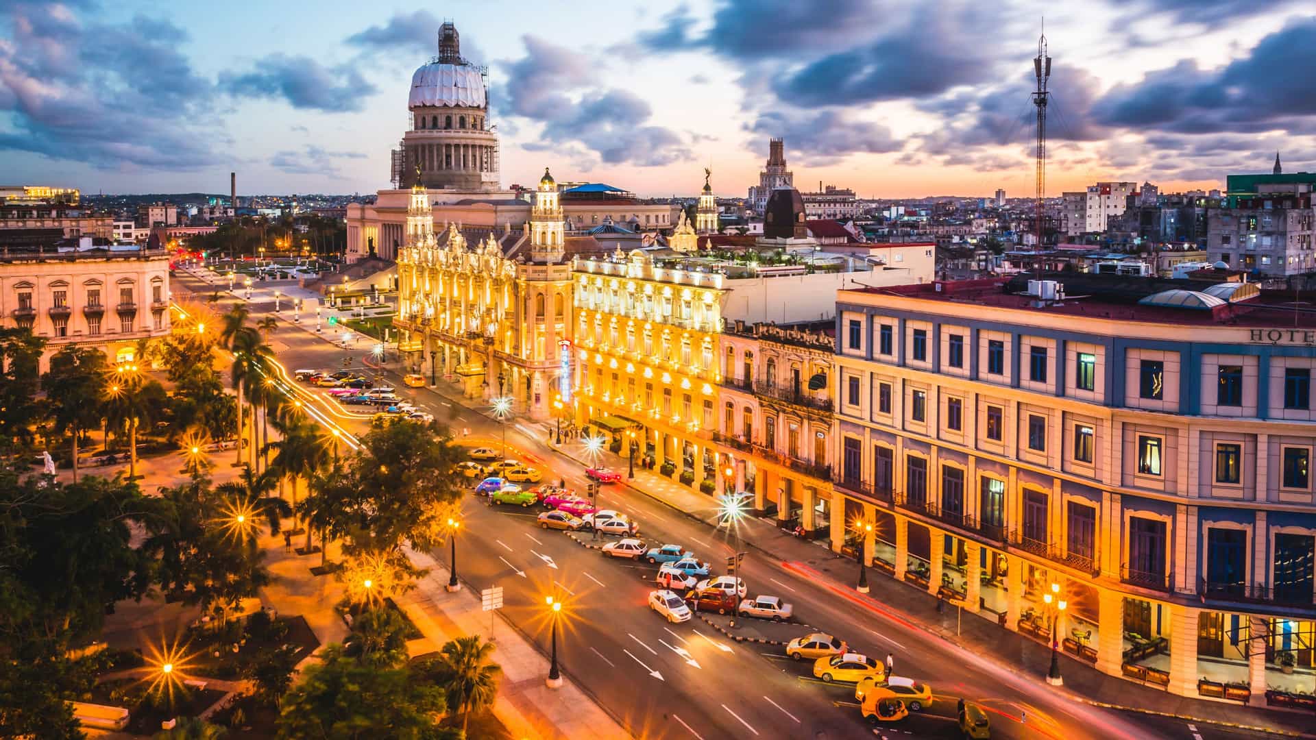 "A wide-angle, elevated view of downtown Havana, Cuba at sunset, showing the iconic Capitolio building, classic cars on the bustling street, and glowing streetlights.  "