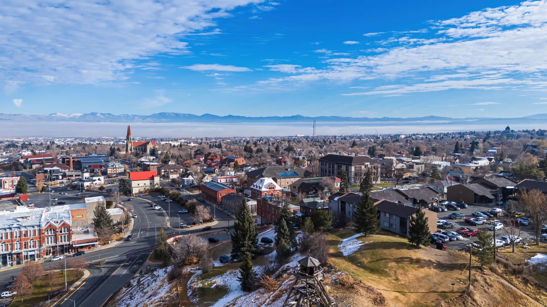 "An aerial view of Helena, Montana, with the city's historic buildings and the iconic Cathedral of St. Helena in the foreground, and the snow-capped mountains of the Rocky Mountains in the distance under a blue sky.  "