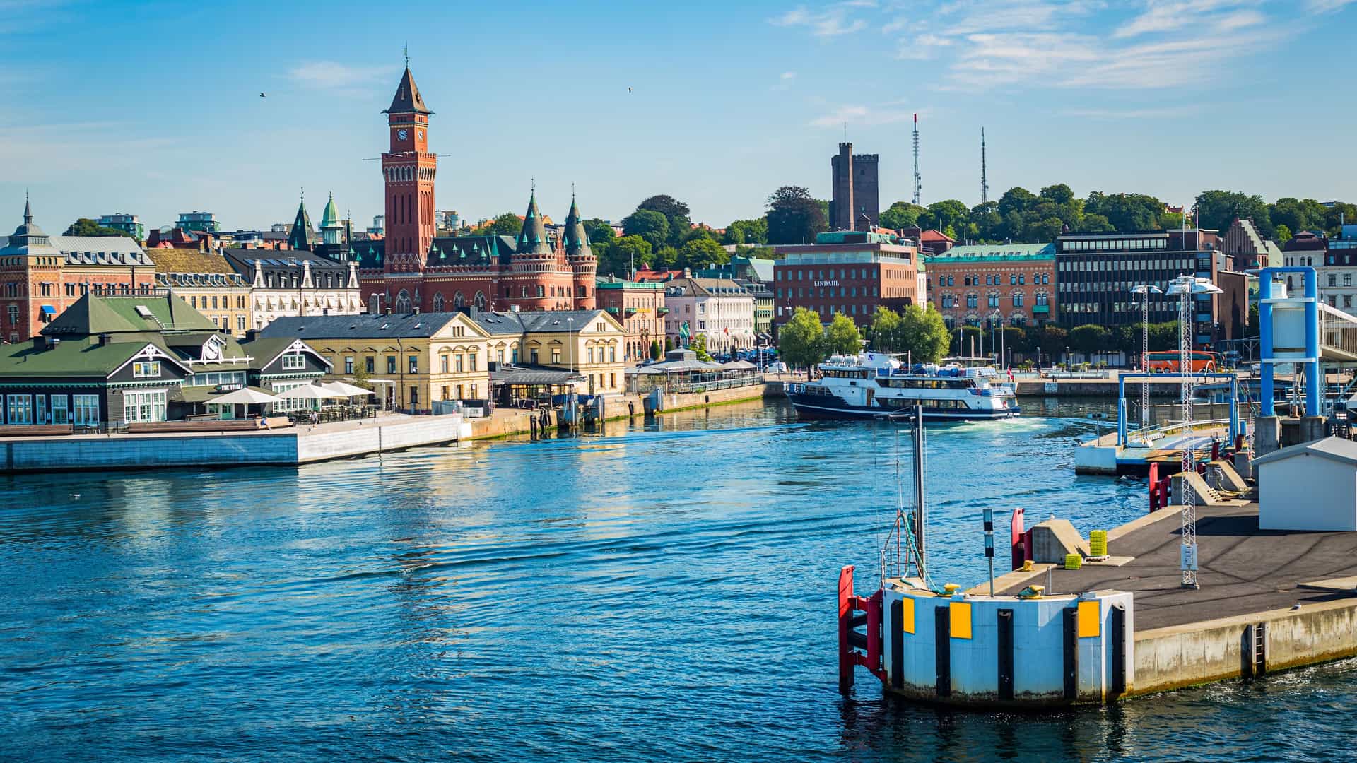 "A sunny panoramic shot of the Helsingborg waterfront in Sweden, with the city's charming architecture and historic clock tower reflected in the harbor's calm, blue water as a ferry approaches the dock.  "