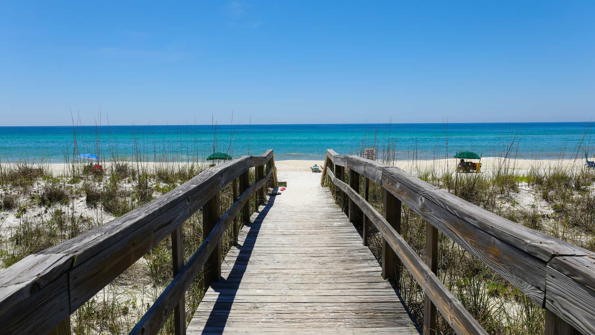 A long wooden boardwalk leads to a pristine white sand beach and the turquoise waters of Henderson Island, with a clear blue sky and gentle waves on the shore.